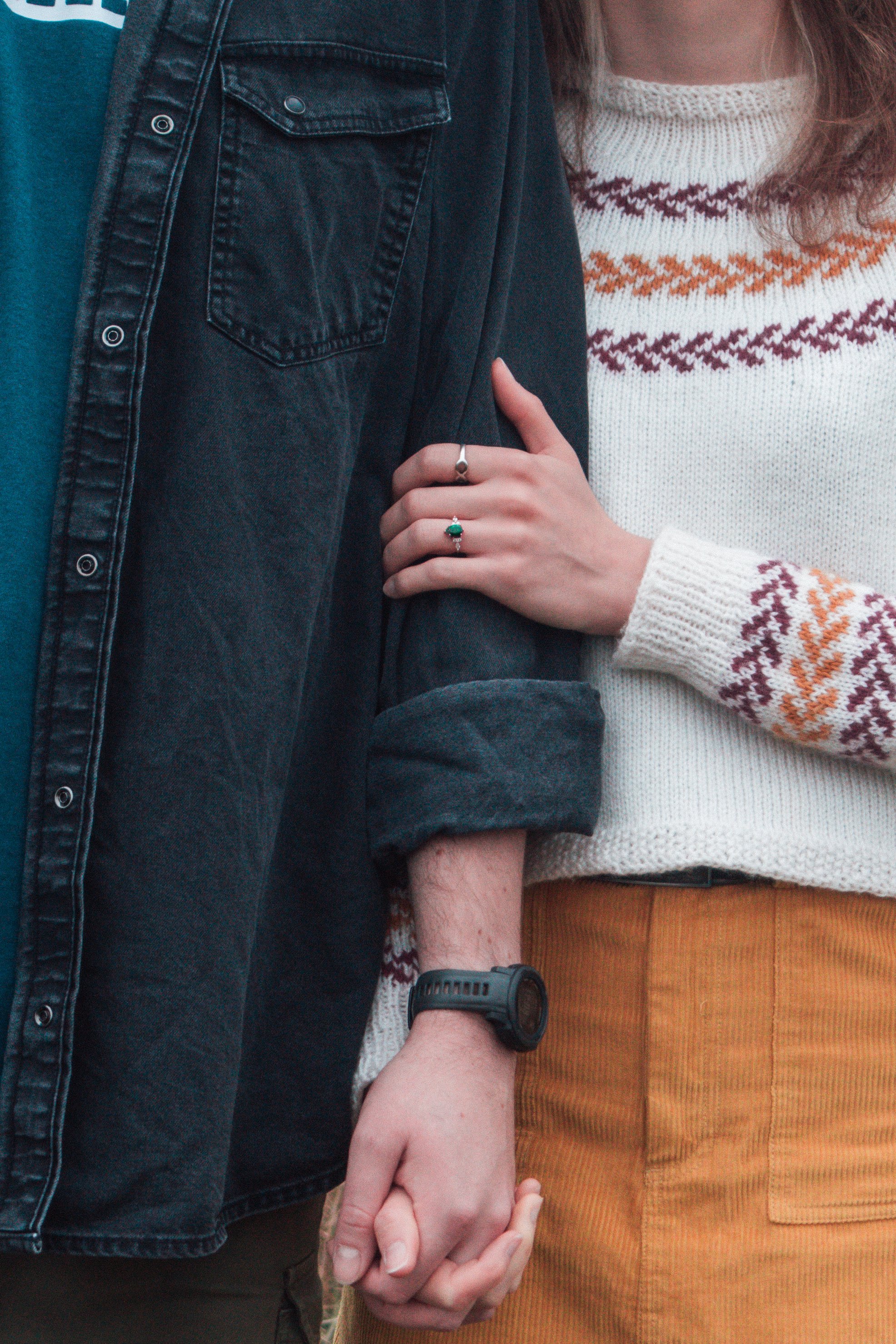 A couple holding hands, with the woman's arm around the man's arm, both wearing casual clothes. The woman is wearing a white sweater with an orange and purple pattern and mustard-colored pants. The man is wearing a black denim jacket and has a black 