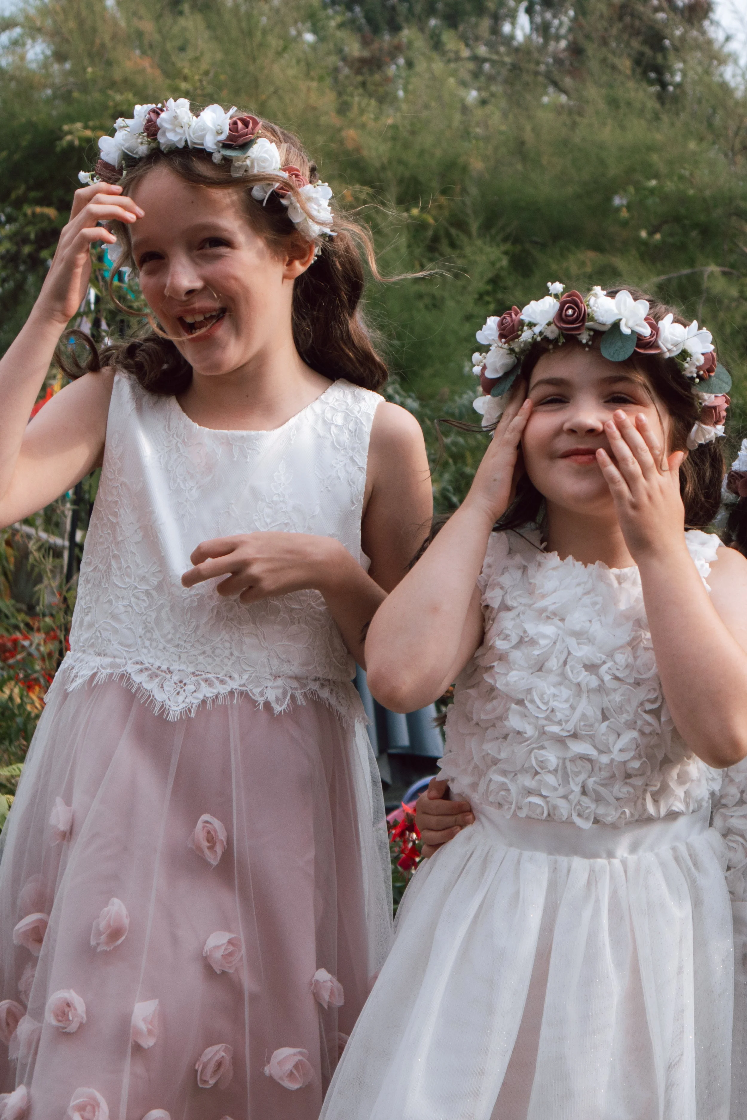 Two young girls wearing floral crowns and dresses, smiling and posing outdoors.