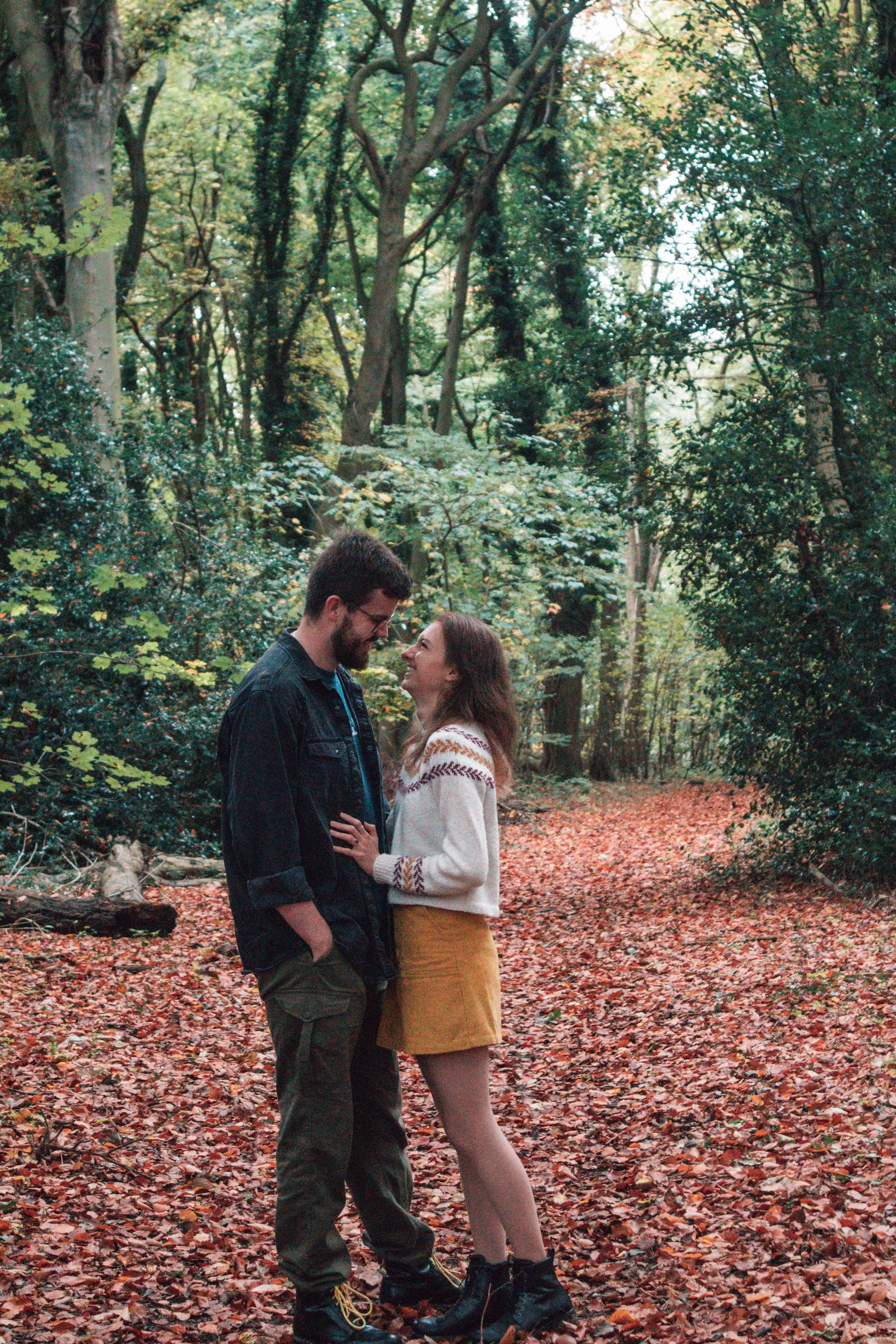 A couple smiling and standing close together on a wooded trail covered in fallen leaves, in an outdoor forest setting during autumn.