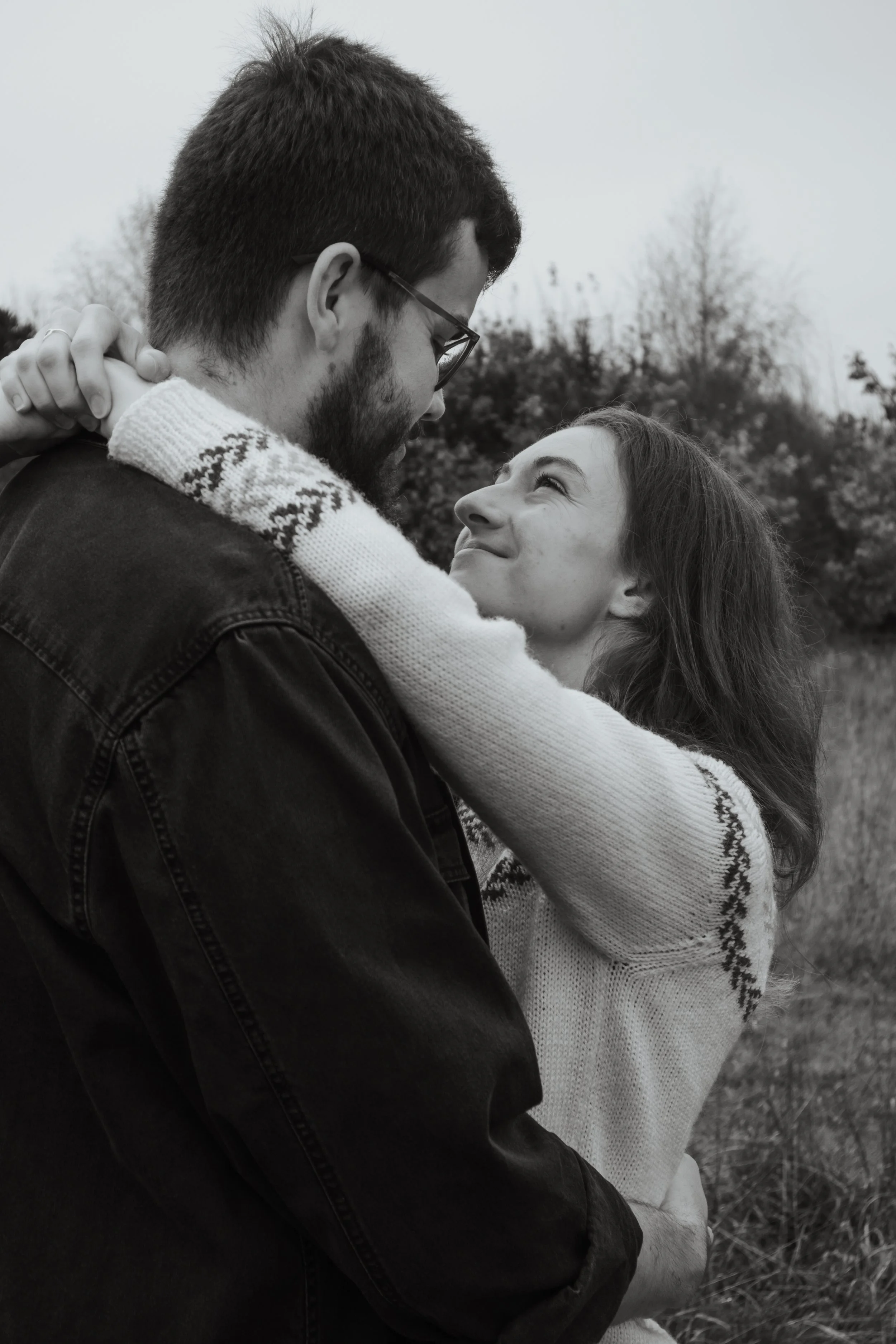 A black and white photo of a couple outdoors, gazing into each other's eyes, with the woman smiling and the man holding her close.