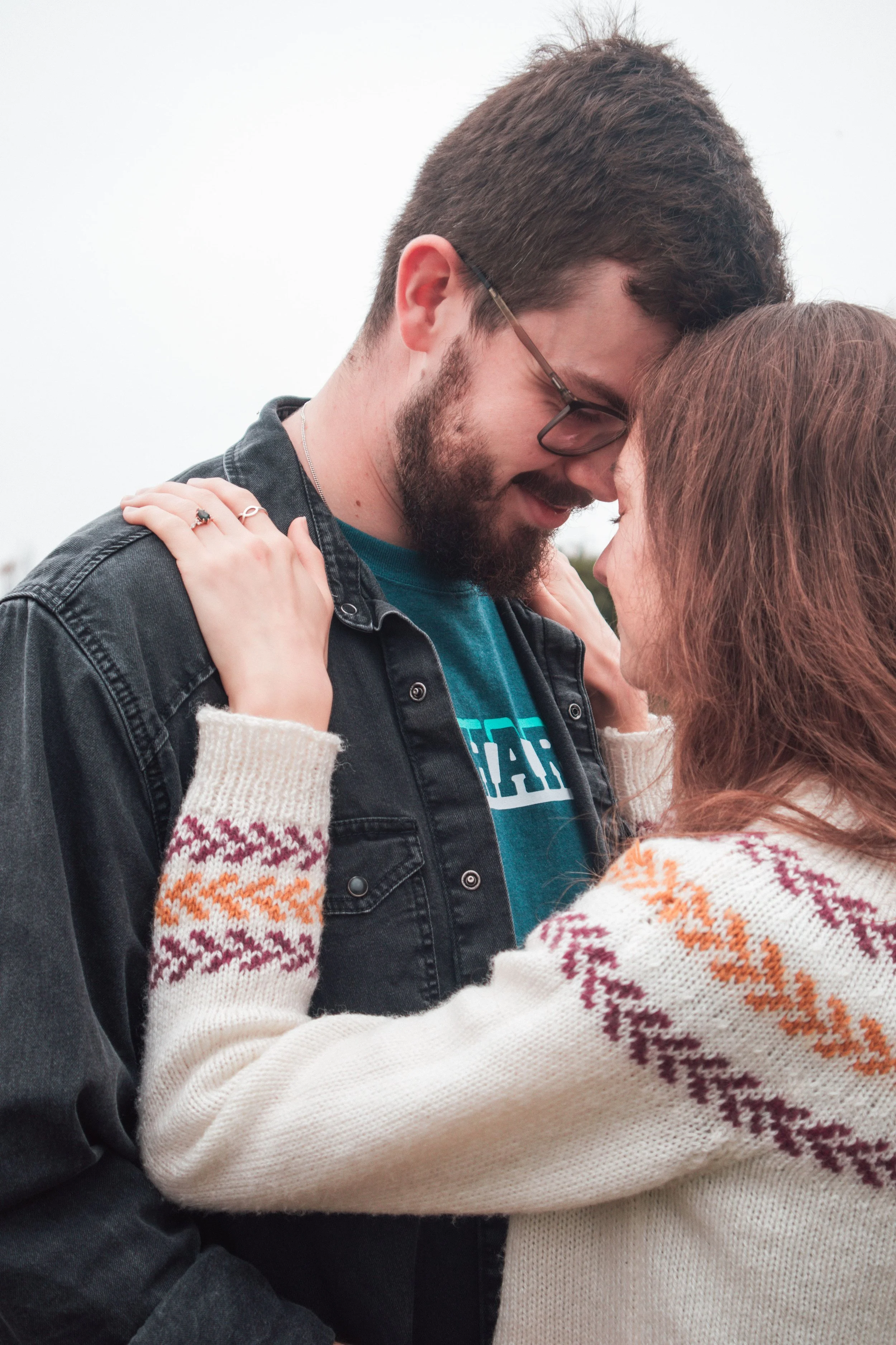 A couple embracing outdoors during overcast weather, touching foreheads and smiling.