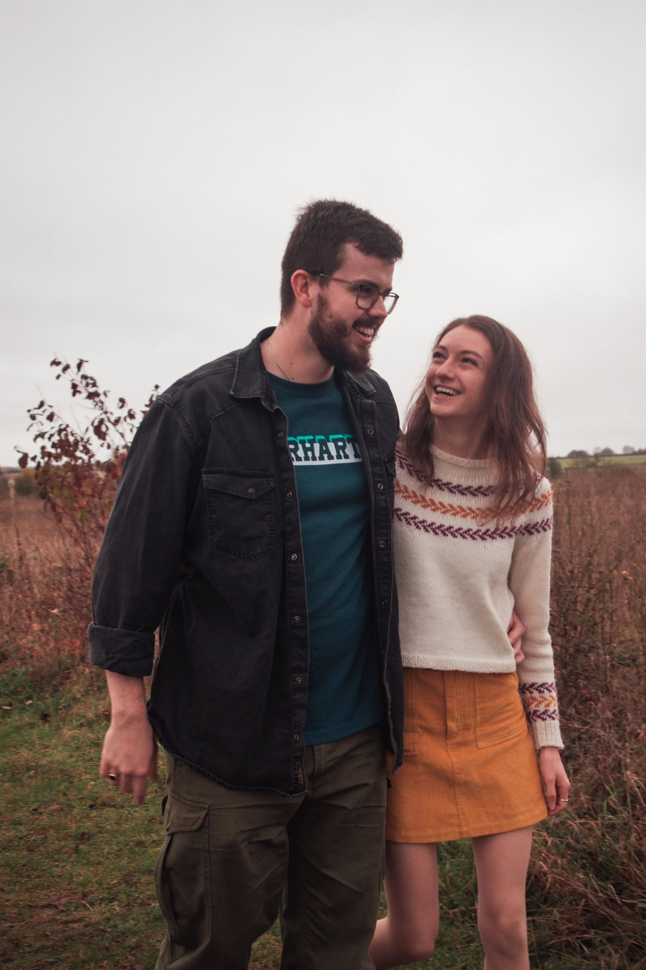 A young man and woman smiling and enjoying each other's company outdoors on a cloudy day, standing in a field with autumn foliage.