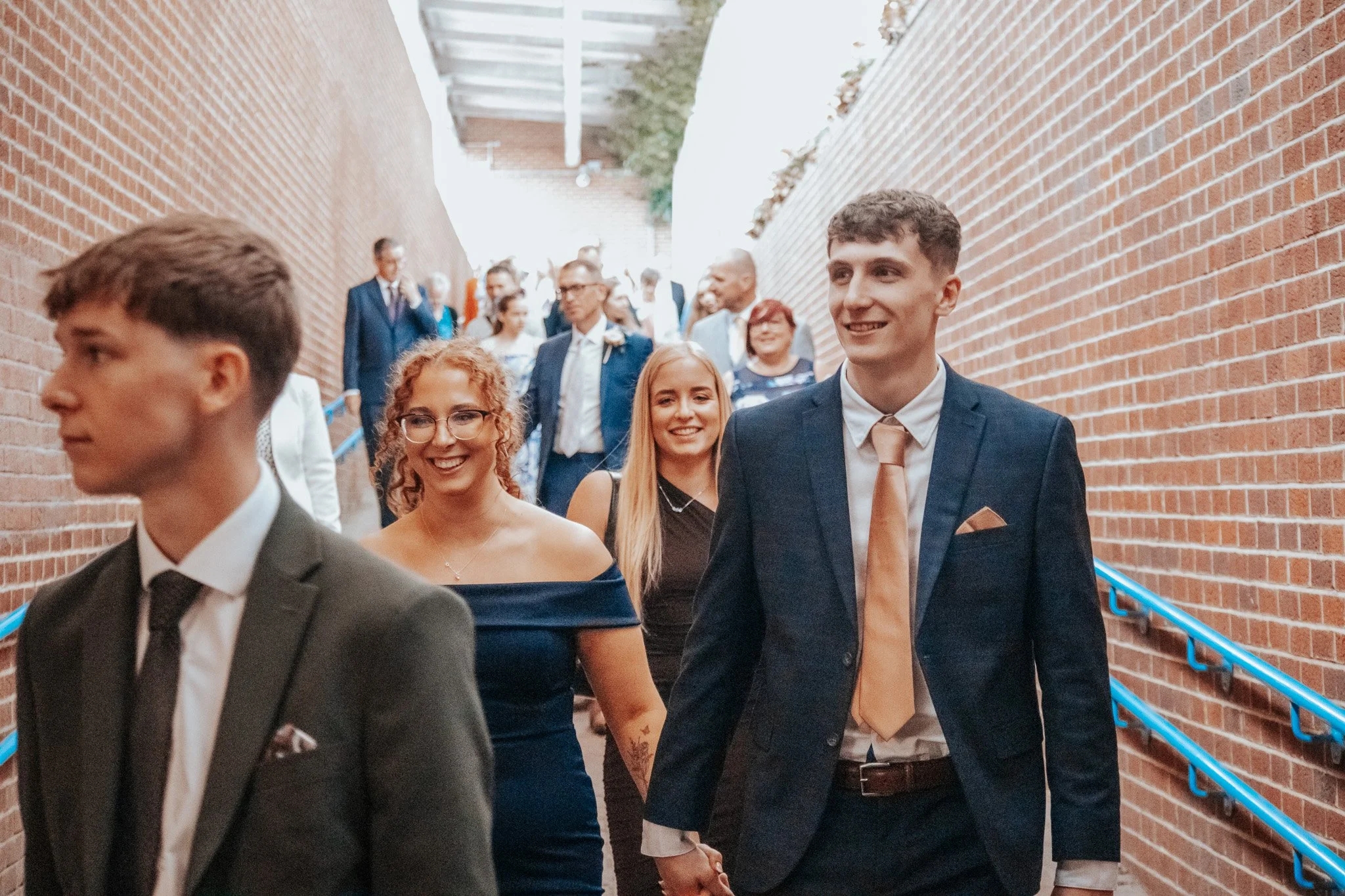 Group of people dressed in formal attire walking through an outdoor corridor with brick walls on either side.
