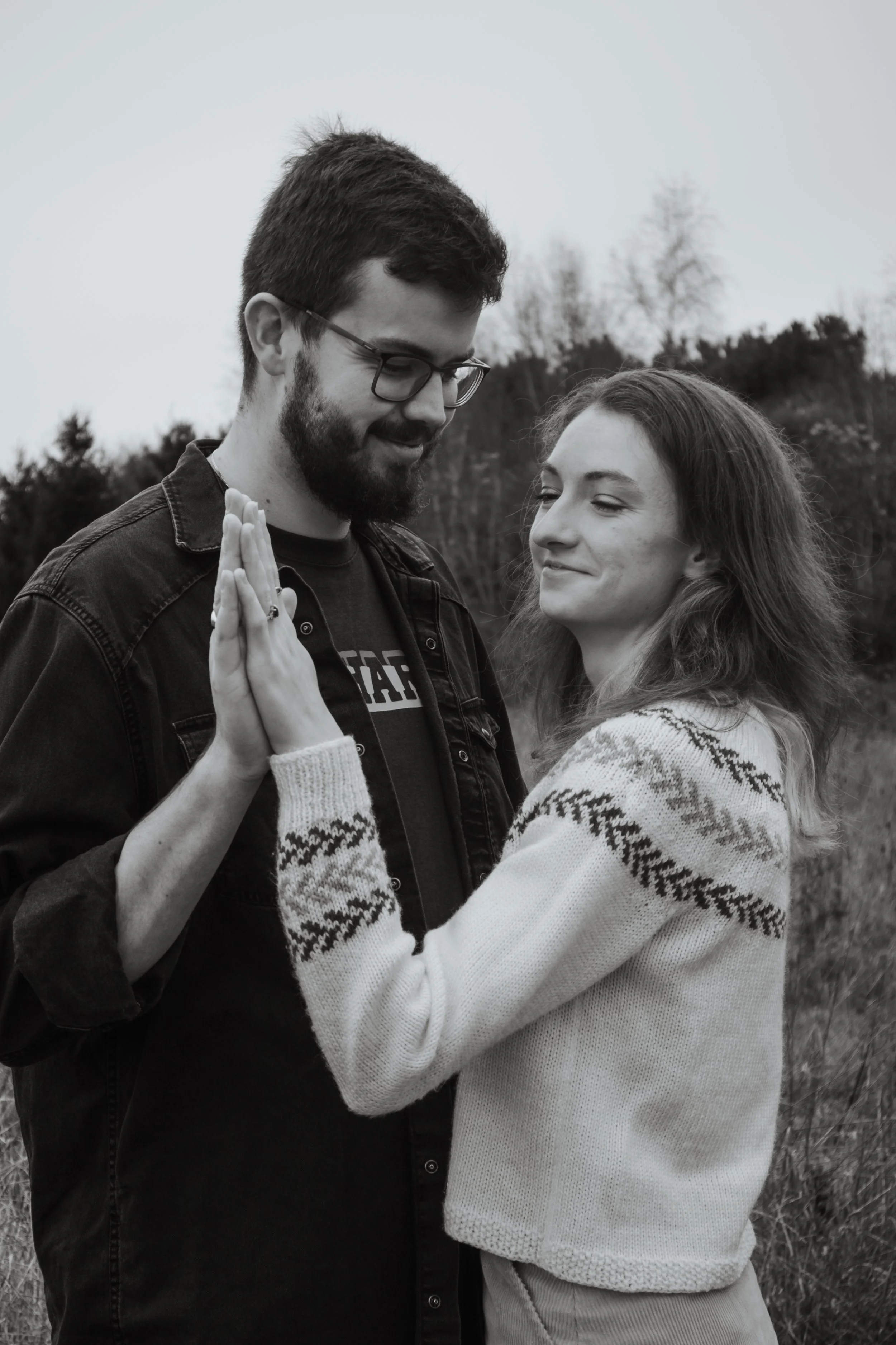 Black and white photo of a couple standing outdoors, touching hands and smiling at each other.