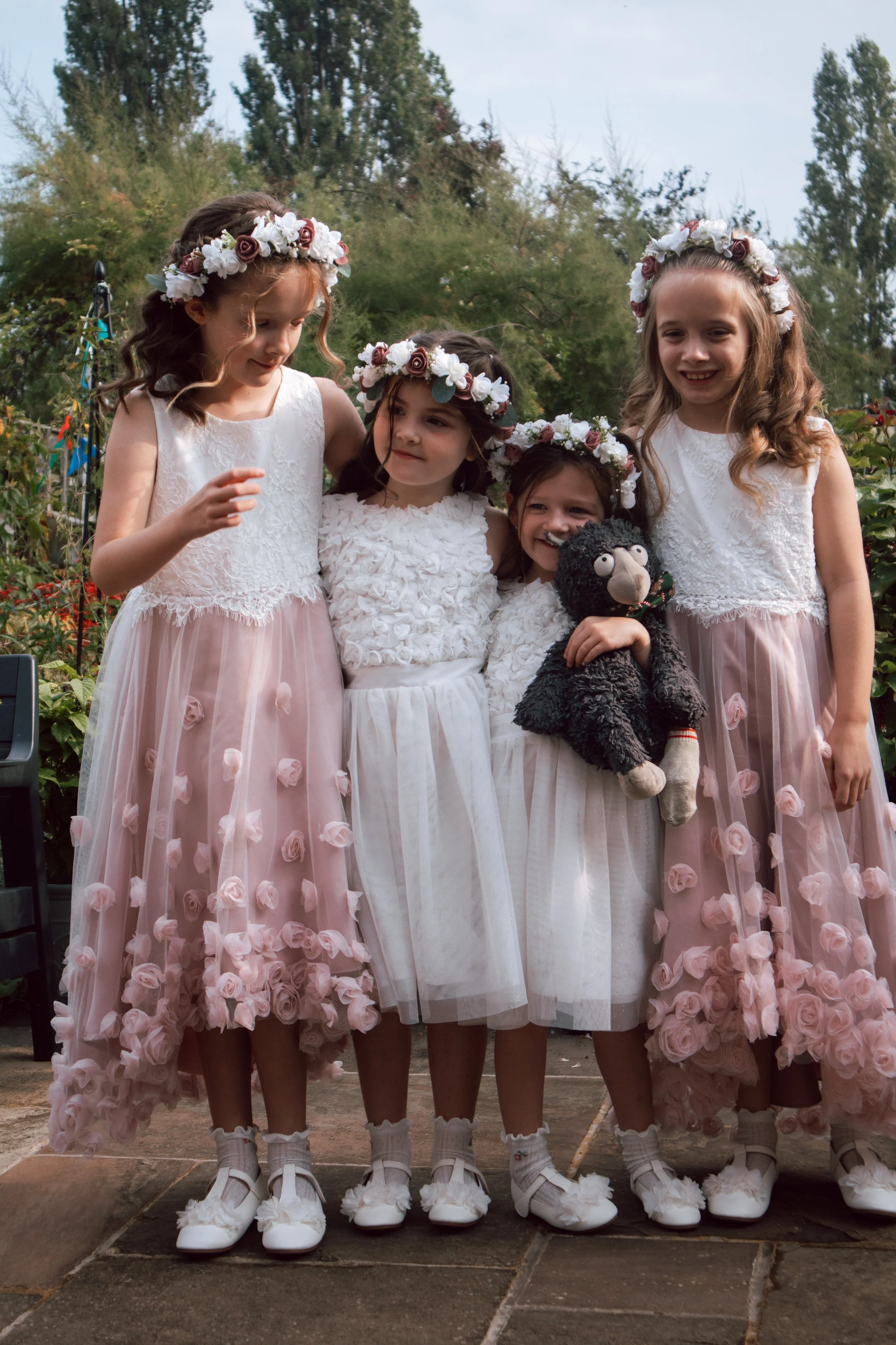 Four young girls wearing white and pink floral dresses and flower crowns, standing together outdoors with greenery in the background, one holding a stuffed animal.