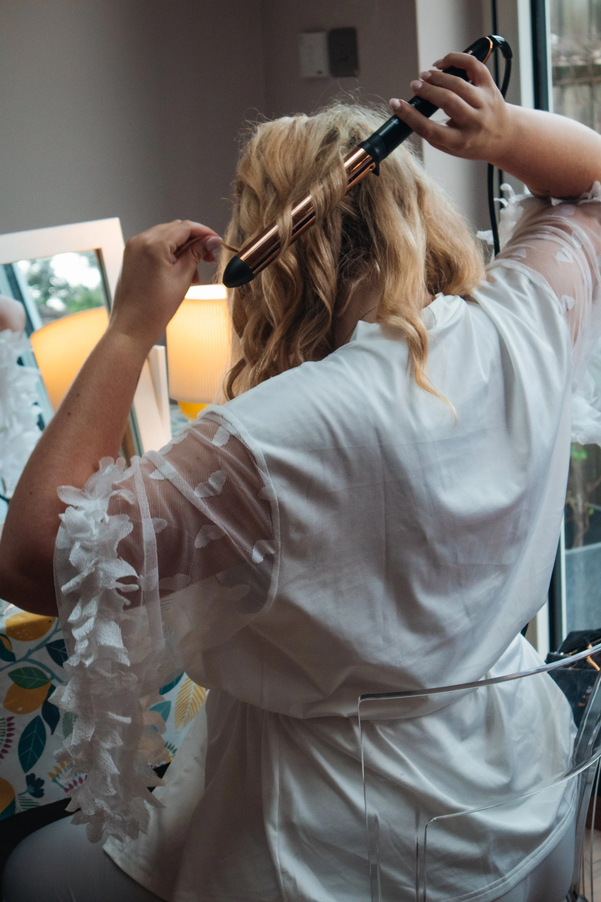 A woman with curly blonde hair is styling her hair with a curling iron in a room with natural light.