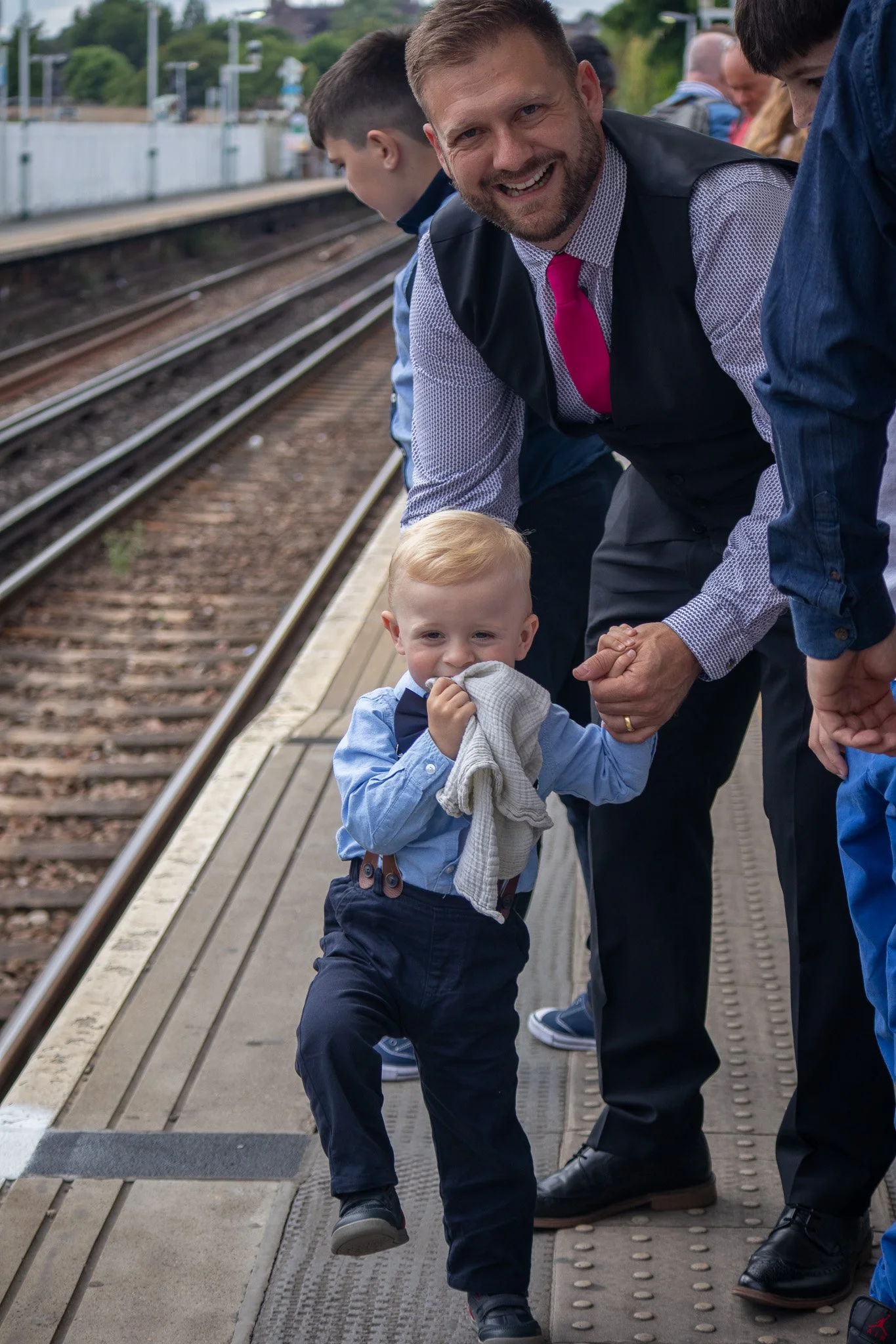 A man holding hands with a young boy on a train station platform, with other children and adults nearby. The boy is holding a cloth in his mouth and smiling.