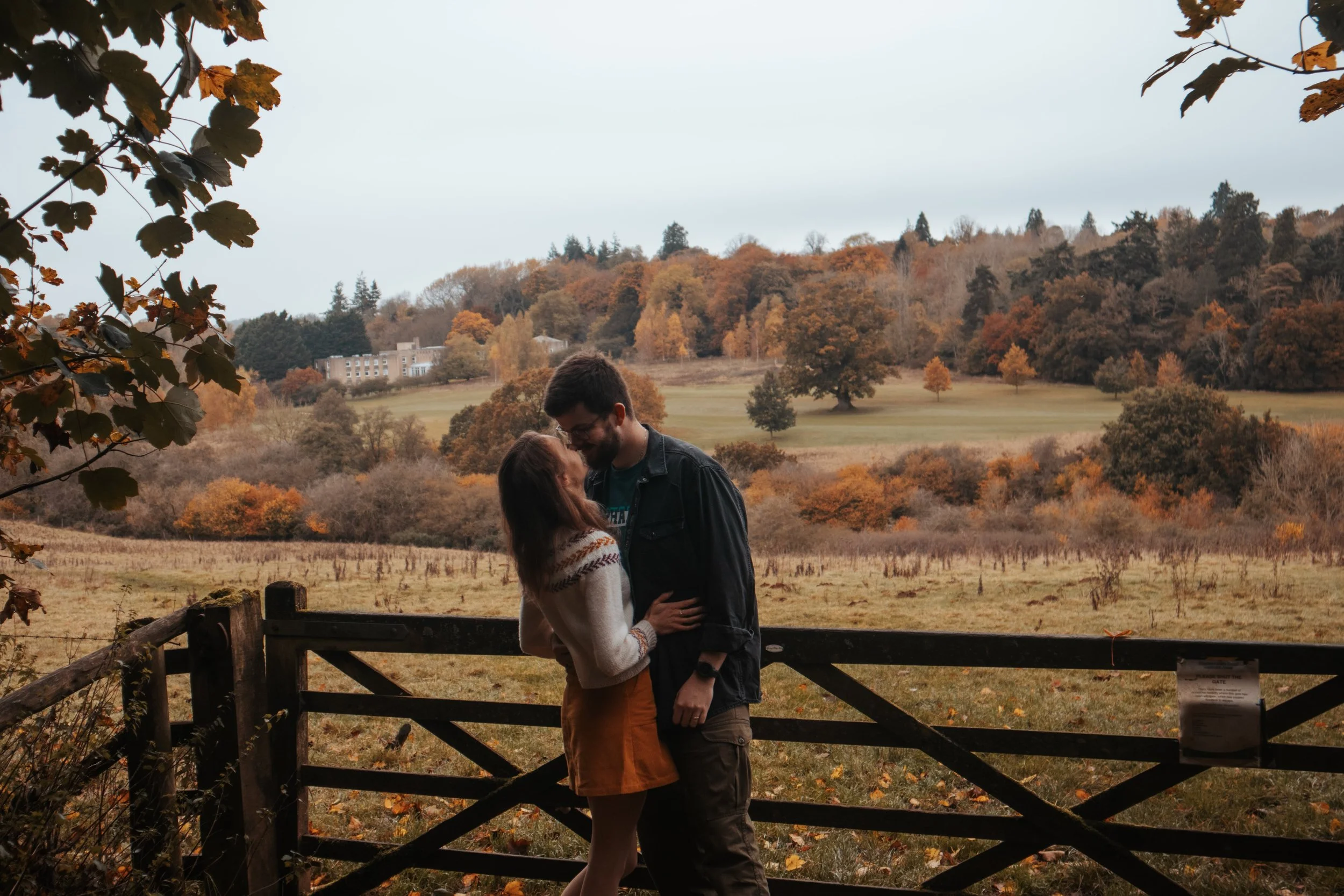 A couple is standing close together, smiling and embracing in front of a wooden fence in an open field during fall. The background features rolling hills and trees with autumn-colored leaves under an overcast sky.