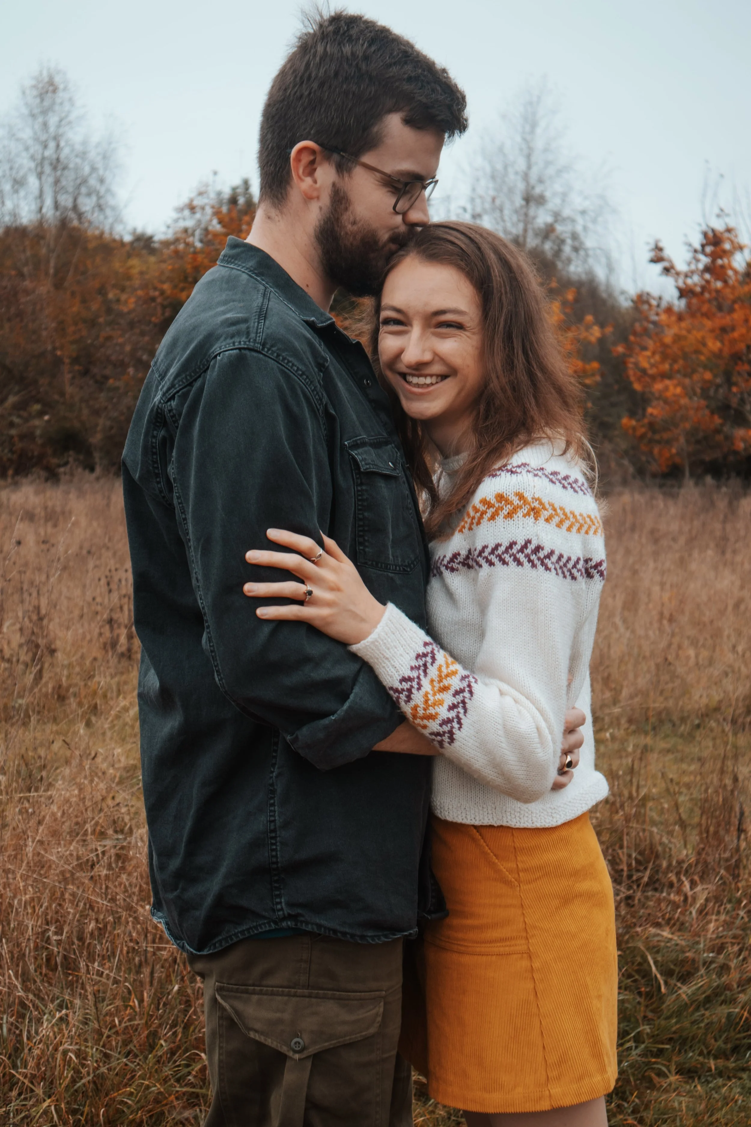 A young couple embracing outdoors in a field with autumn trees, the man is kissing the woman's forehead, both smiling.
