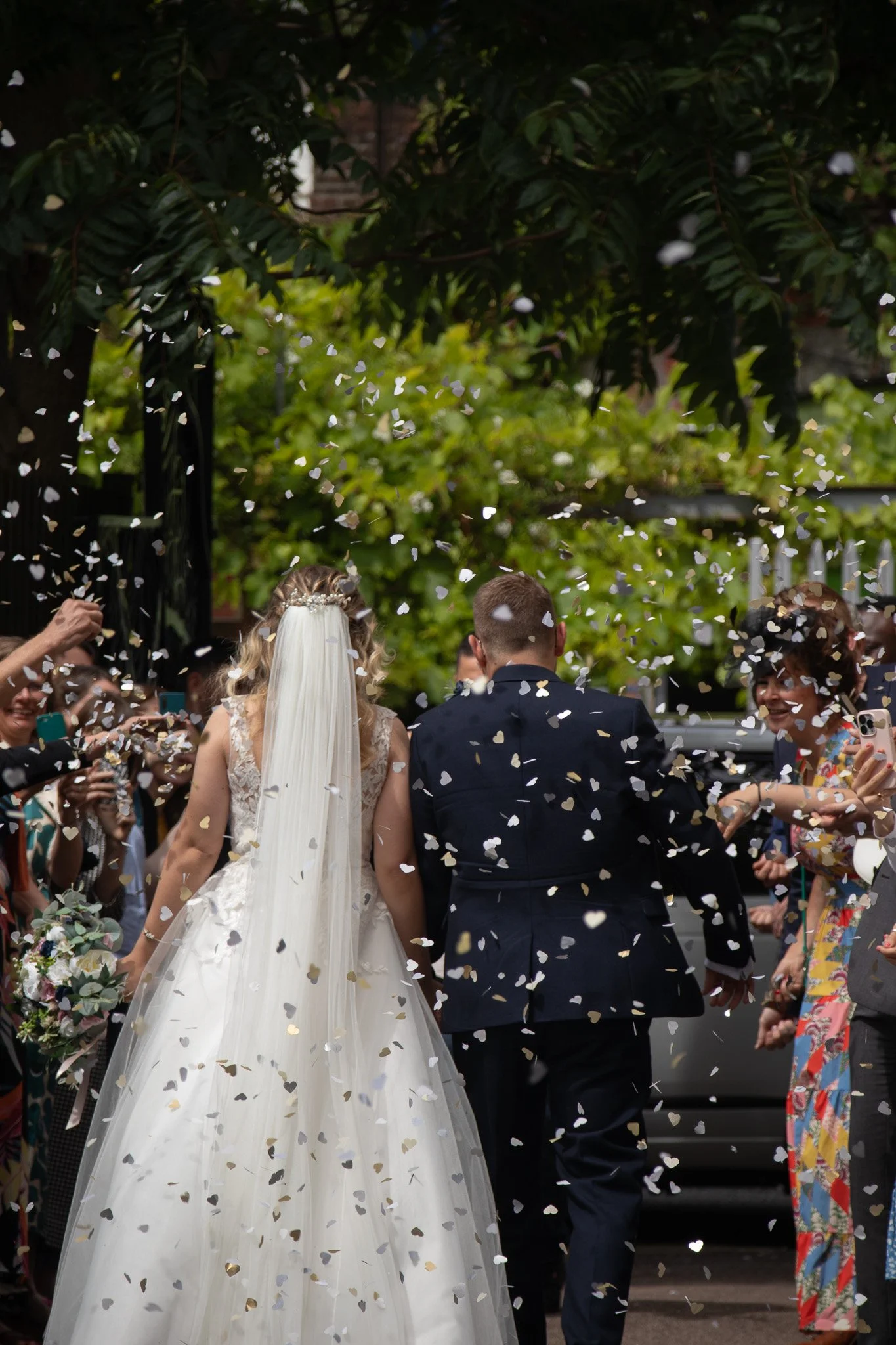 Bride and groom walking through a shower of white confetti, surrounded by friends and family taking pictures and celebrating outside.