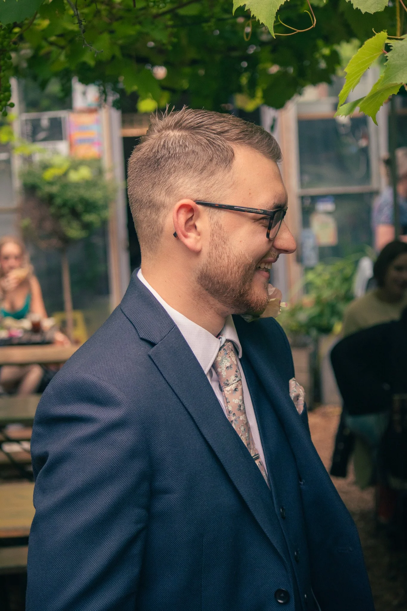 A man with glasses, a beard, and a short hairstyle, smiling while wearing a dark blue suit, white shirt, and a patterned tie, outdoors under green foliage at a social gathering.