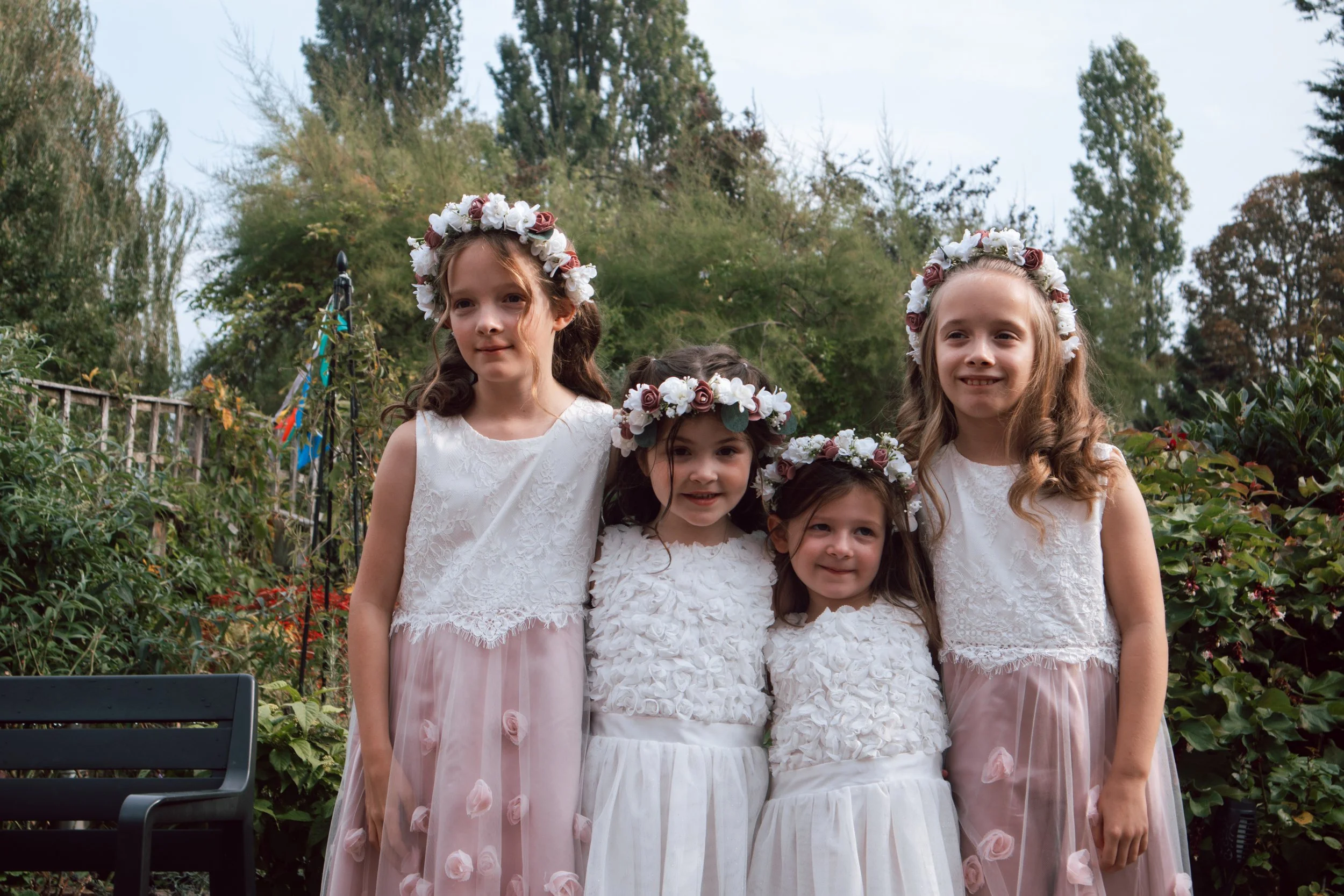 Five young girls in white dresses with floral crowns standing outdoors in a garden.