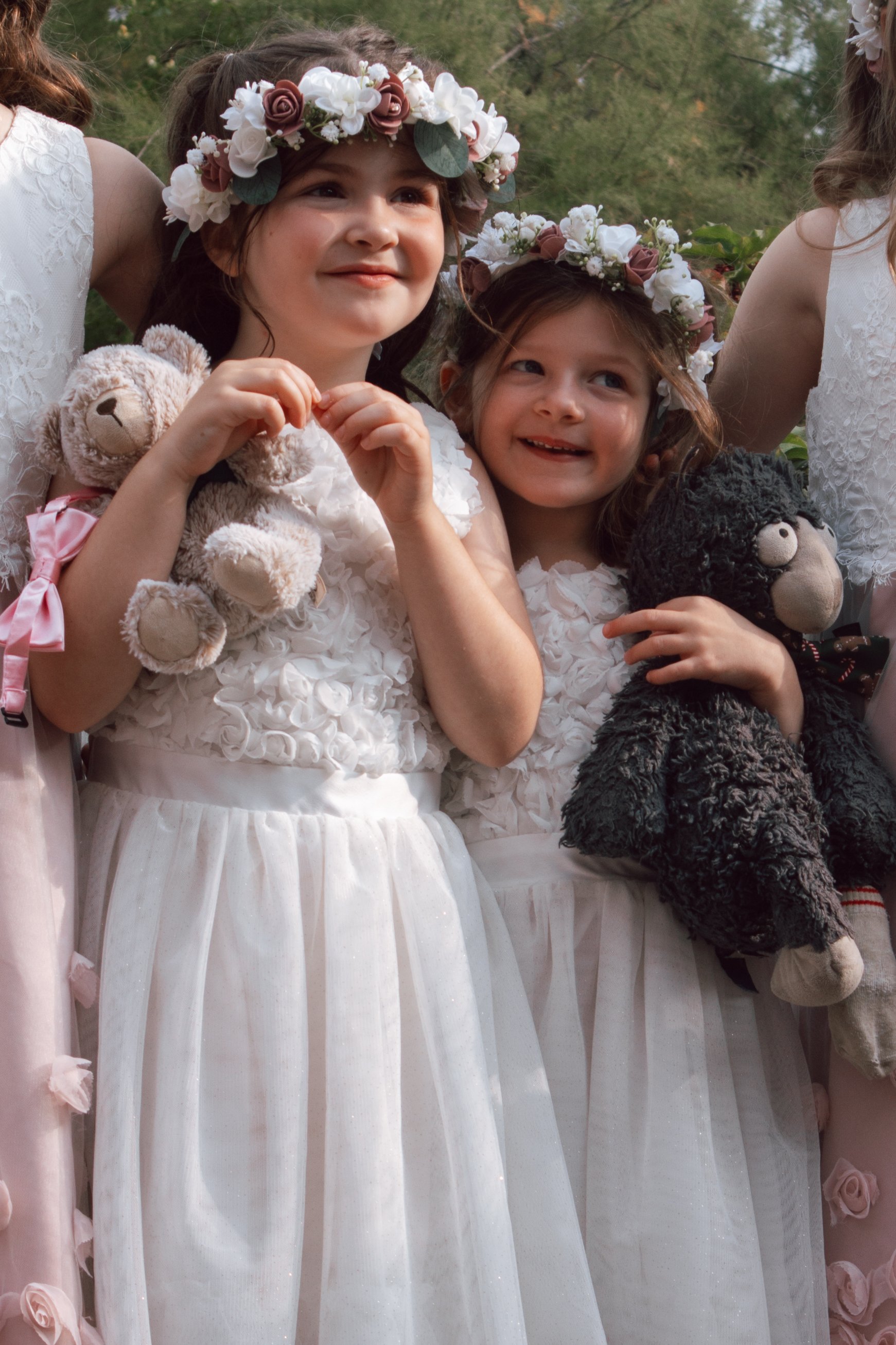 Two young girls wearing white dresses and floral crowns, holding teddy bears, smiling outdoors.