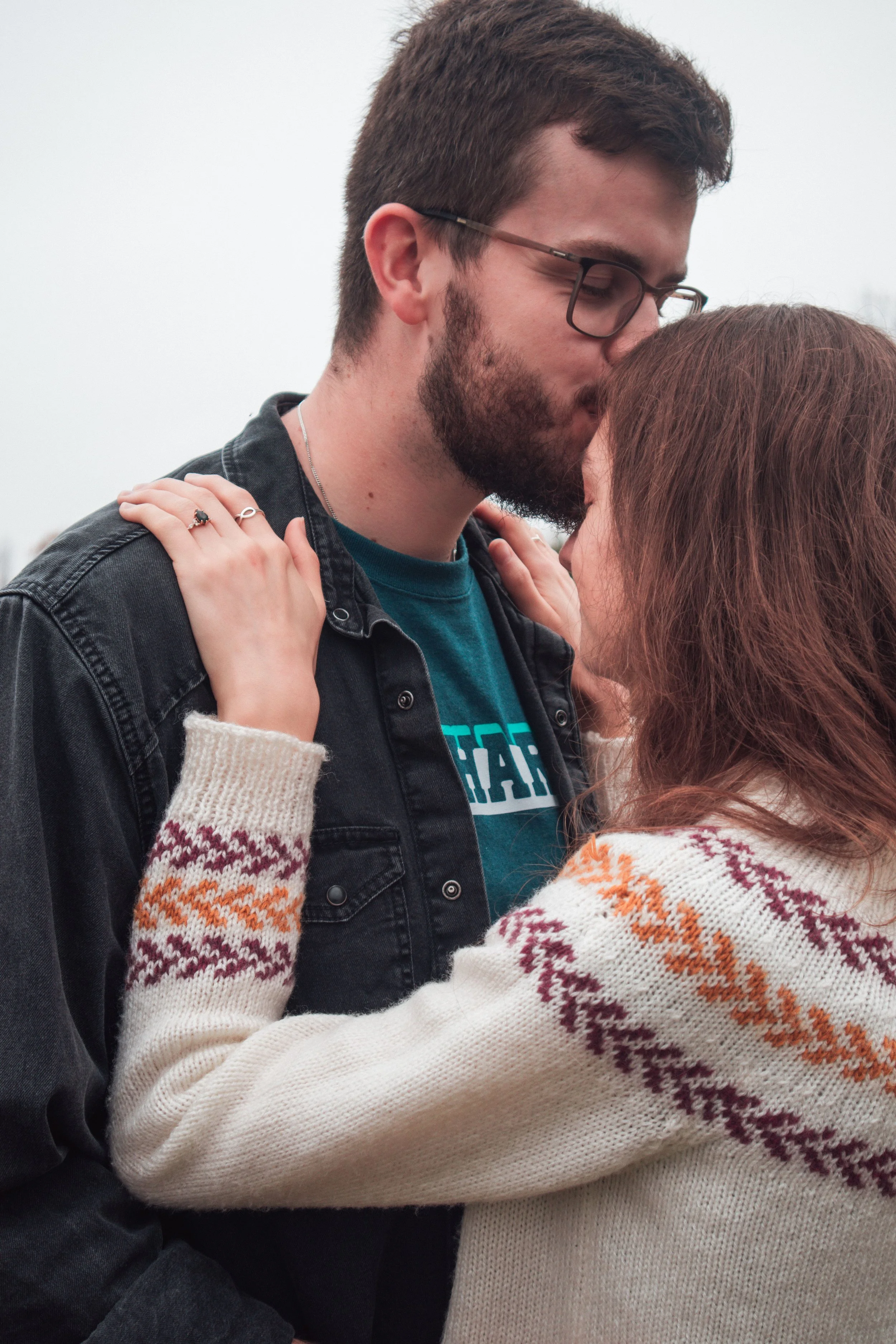 A couple is embracing outdoors, with the man kissing the woman's forehead. The man has a beard, glasses, and is wearing a dark denim jacket over a blue T-shirt. The woman has brown hair and is wearing a cream-colored sweater with colorful patterns on
