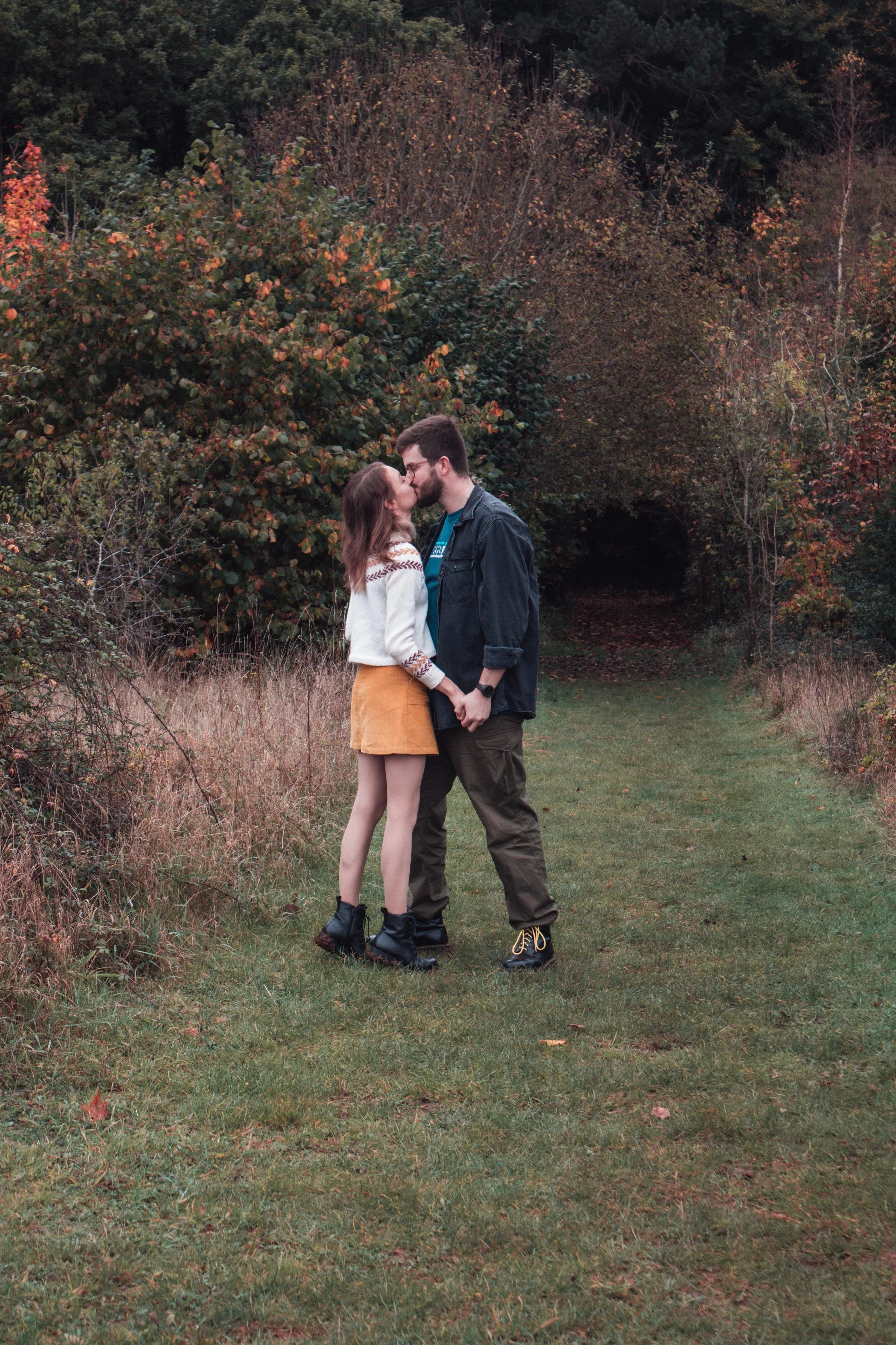 A young couple sharing a kiss in a wooded outdoor area during autumn, holding hands while standing on a grassy path surrounded by fall foliage.