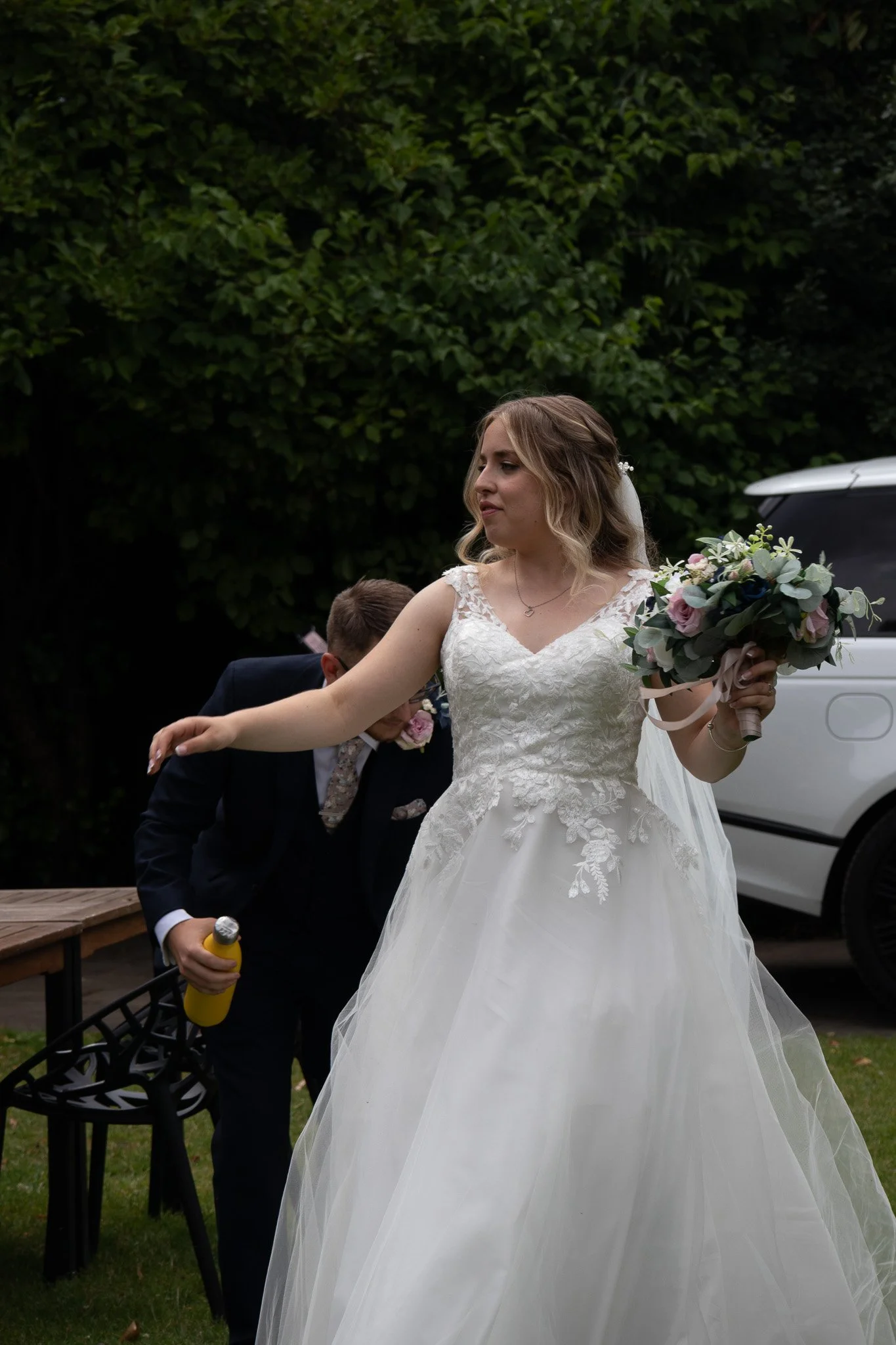 A bride in a white wedding dress holding a bouquet of flowers, standing outdoors near a black chair and a white car, with greenery in the background.