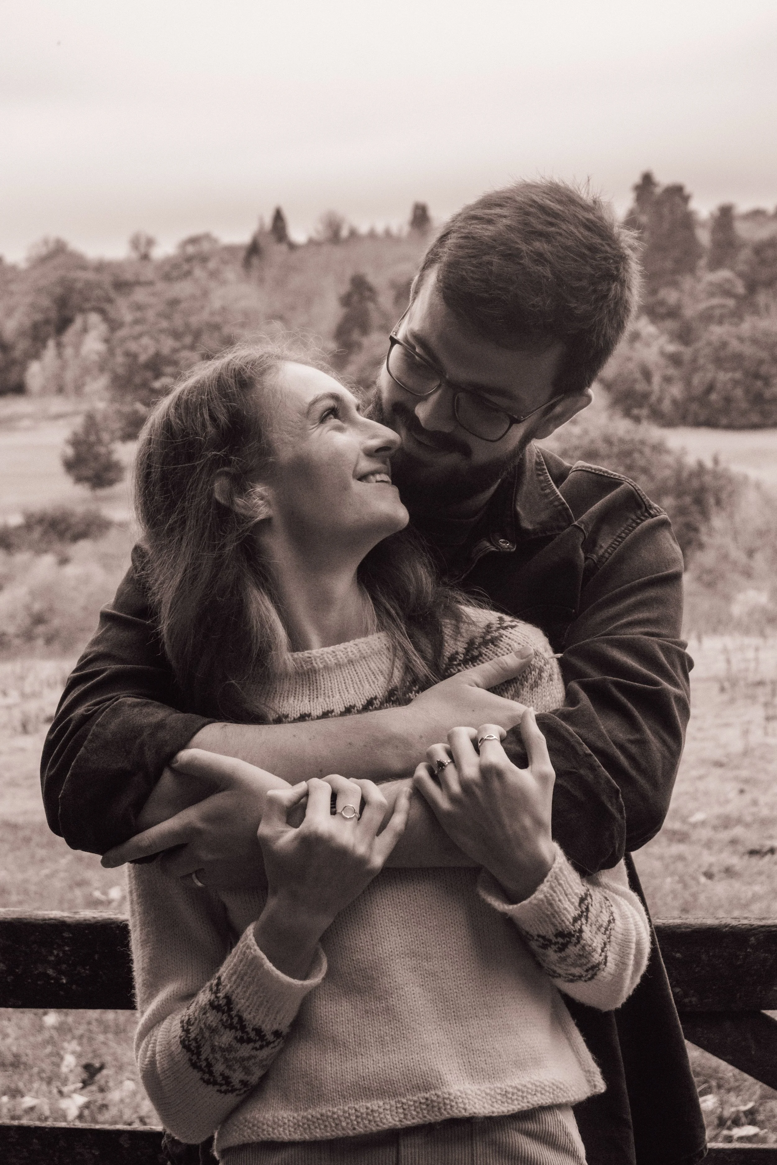 A black and white photo of a couple hugging outdoors, with the woman smiling up at the man and the man looking down at her.