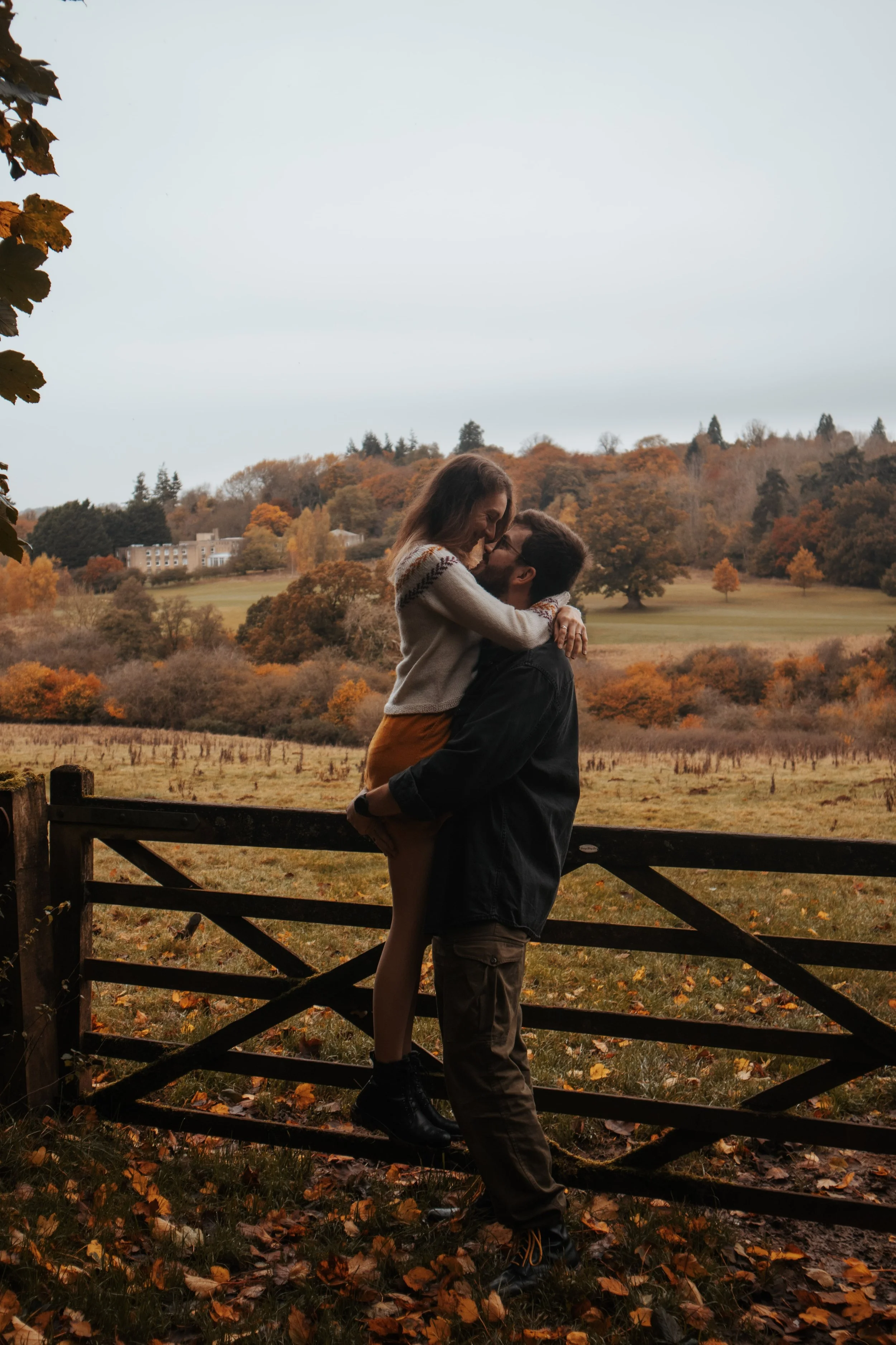 A couple sharing a kiss outdoors on a fall day, with trees in autumn colors and a wooden gate in the background.