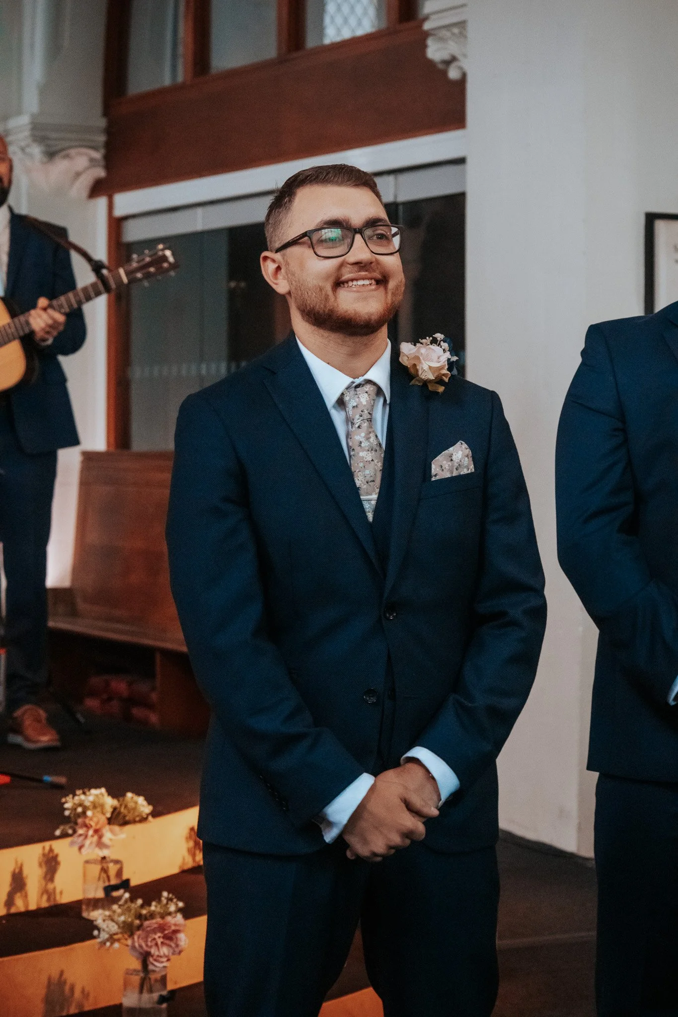 A man dressed in a dark blue suit, white shirt, and patterned tie, smiling at a wedding ceremony, with a flower boutonniere on his lapel. In the background, a musician playing guitar is visible.
