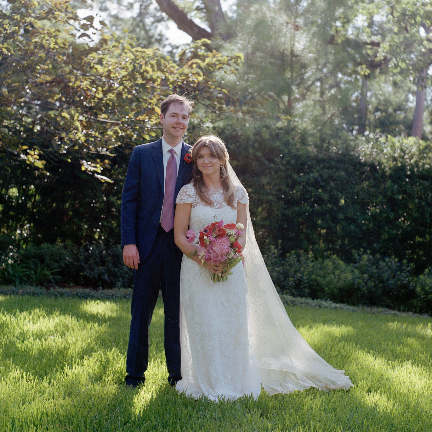 A bride and groom stand on a green lawn in a park, smiling at the camera with trees and bushes in the background. The bride wears a white lace wedding dress and holds a pink and red floral bouquet. The groom wears a navy suit with a pink tie and a re
