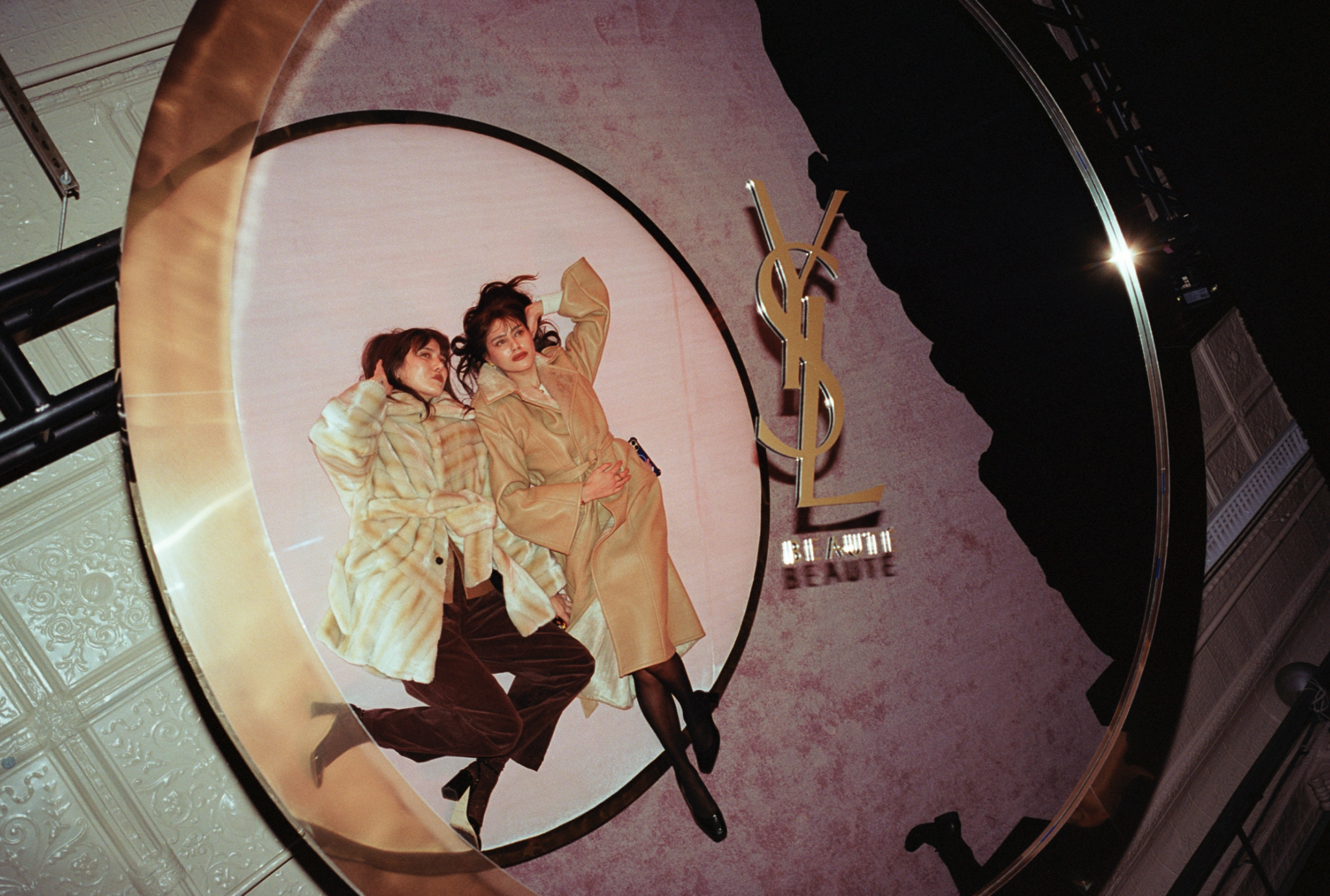 Two women pose on a circular platform with a YSL logo. One woman with dark hair, wearing a beige coat and black shoes; the other with brown hair, wearing a plaid coat and brown pants. The background features a pinkish wall with a sign that says 'BEAU