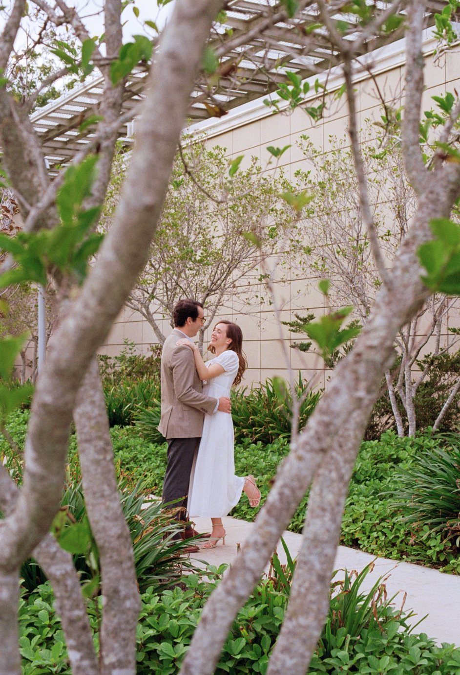 A couple dancing outdoors on a pathway surrounded by green bushes and trees, with a beige building in the background, seen through branches.