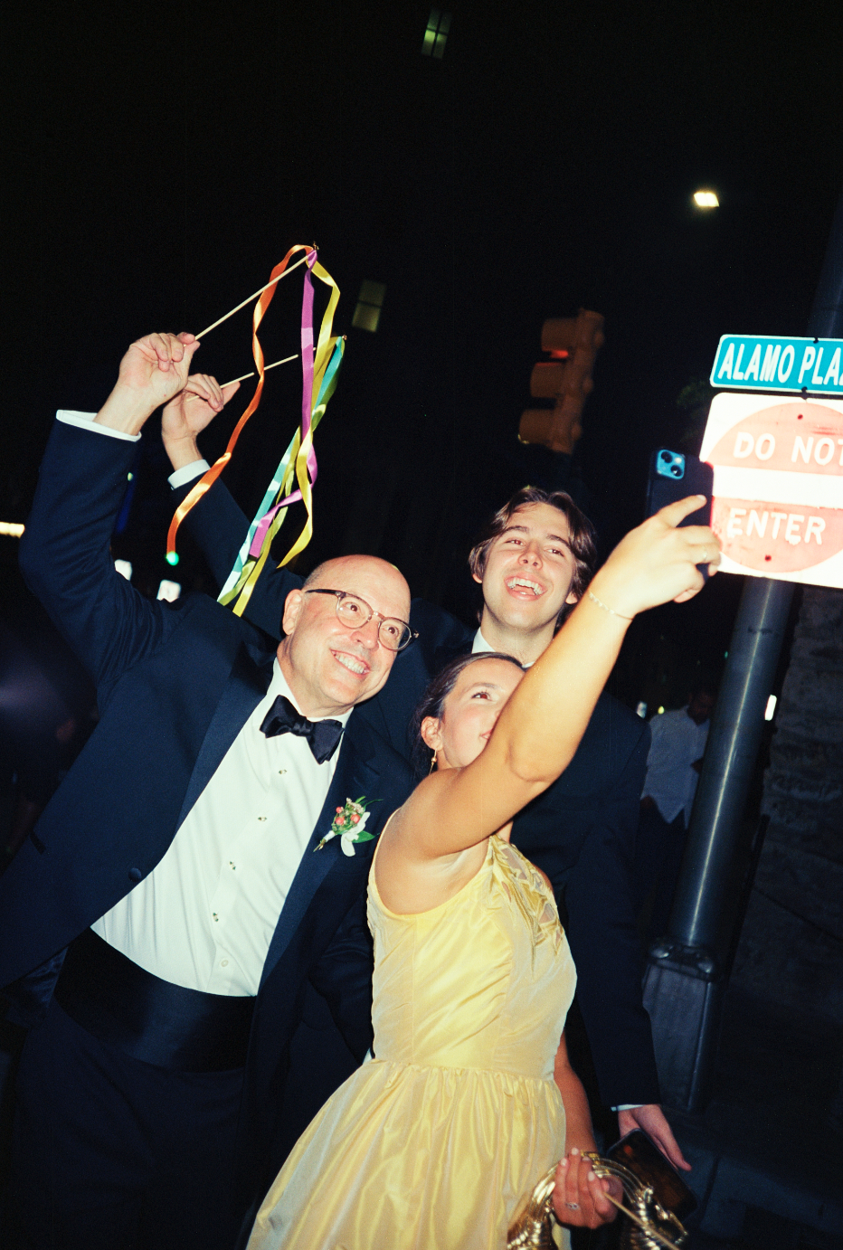 People celebrating outdoors at night, taking a selfie, with a street sign reading 'Alamo Plaza' and a 'Do Not Enter' sign in the background.