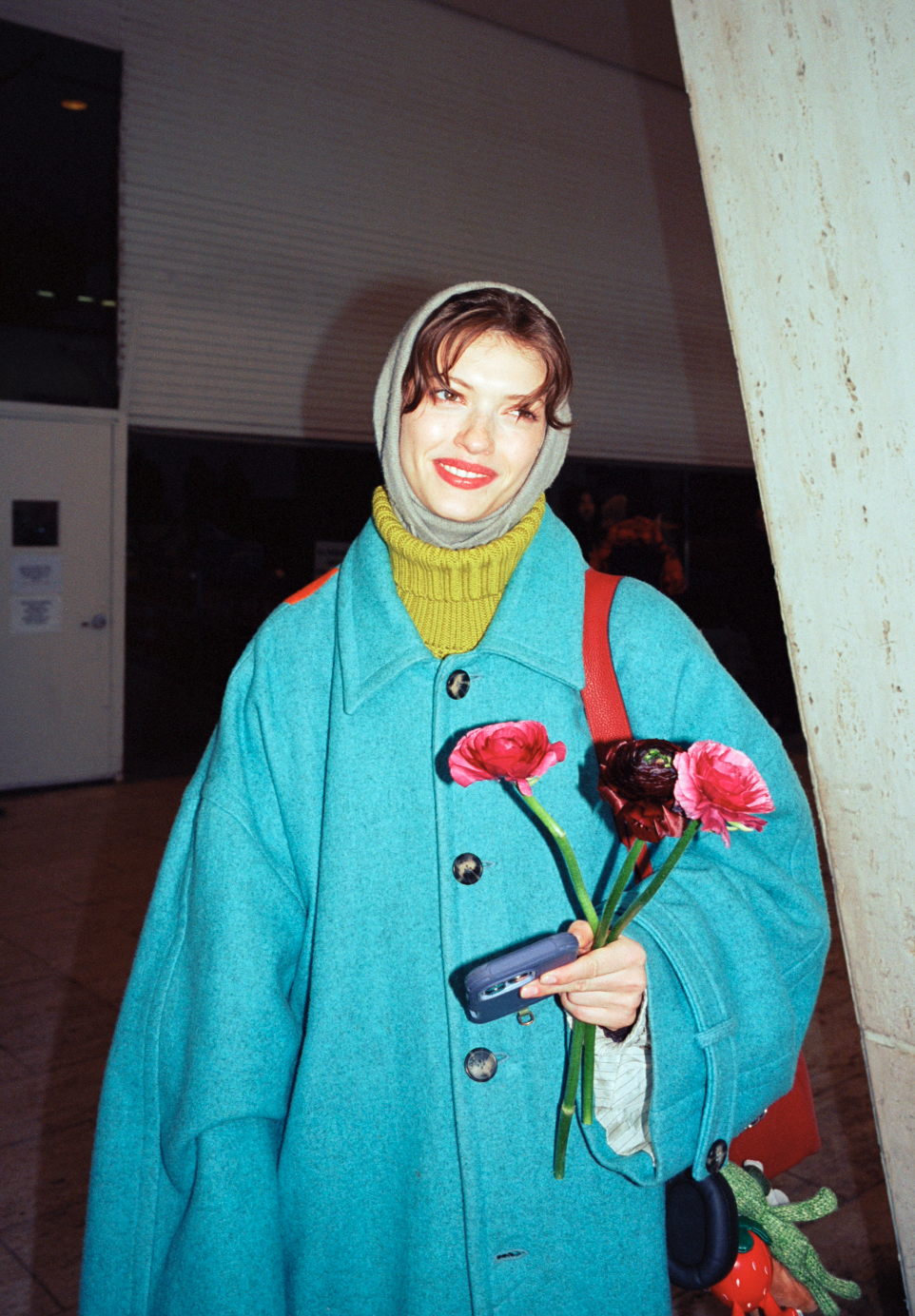 A woman wearing a gray hood, a bright blue coat, a yellow-green scarf, and red lipstick holding a small bouquet of pink and purple flowers in her left hand and a cellphone in her right hand. She is smiling and standing indoors near a large concrete p