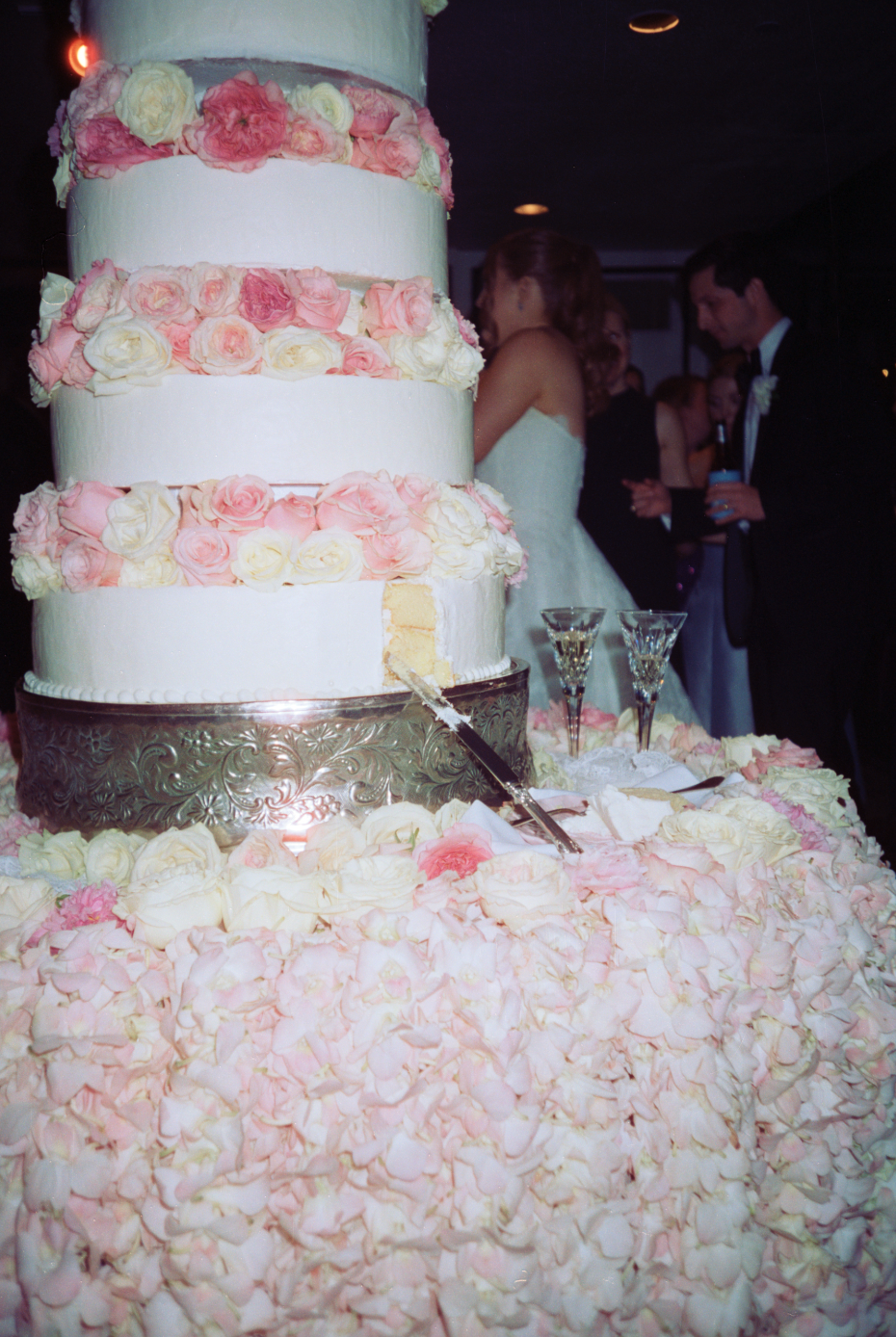 A large, multi-tiered wedding cake decorated with pink and white roses on a table covered with pink and white rose petals. In the background, a bride in a white wedding dress and a groom in a black tuxedo are seen, along with wedding guests at a cele