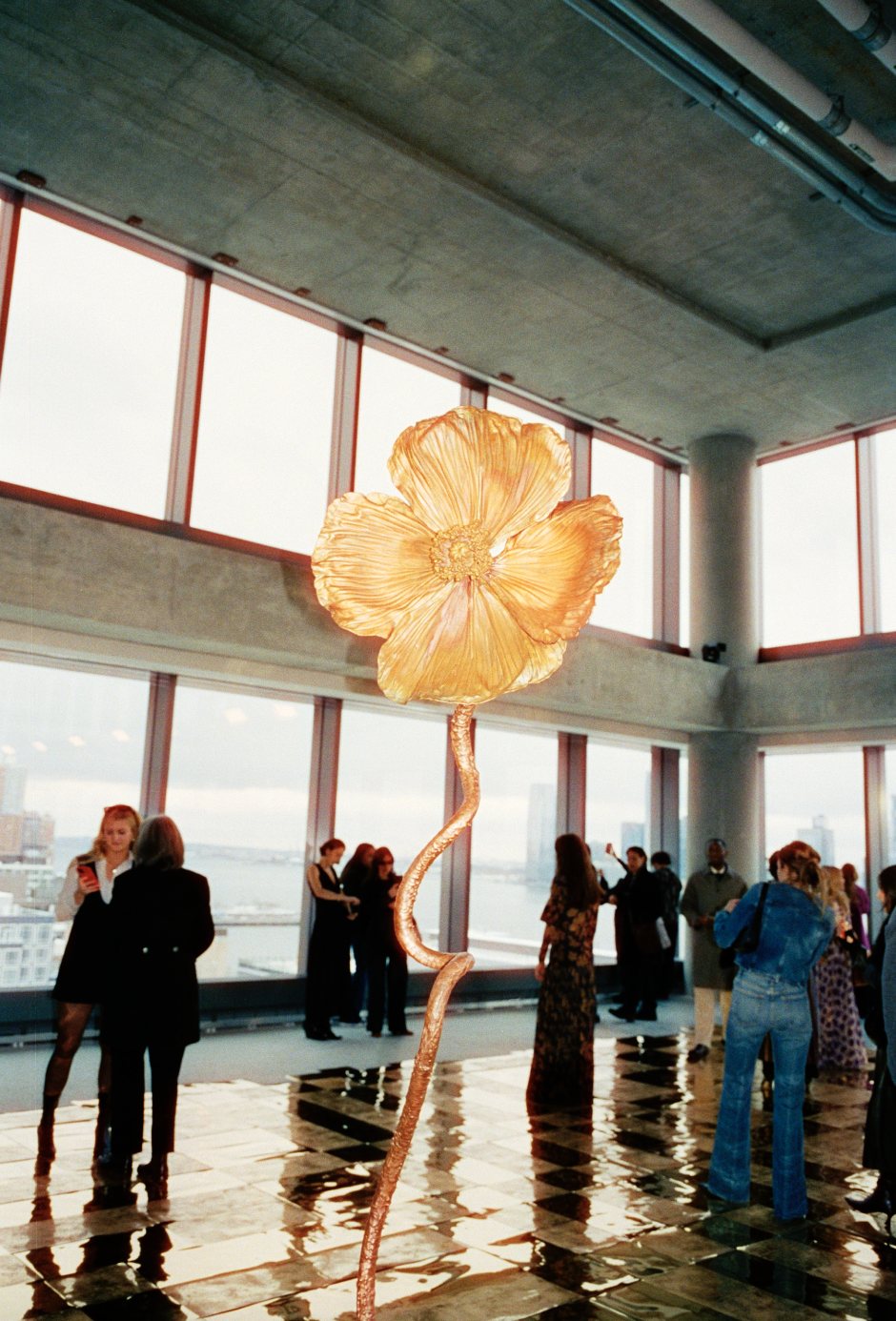 An art installation of a large, orange flower with a wavy stem inside a modern office or gallery space with large windows and several people in the background.