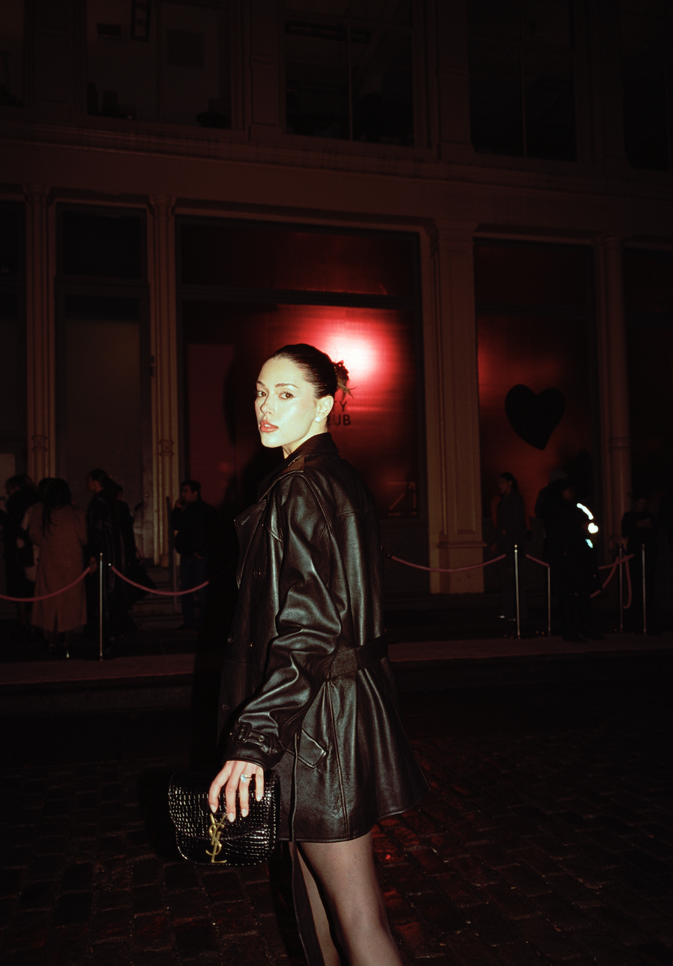 Woman in a leather coat holding a YSL clutch purse standing on cobblestone street at night, with a club entrance and other people in the background illuminated by red lights.