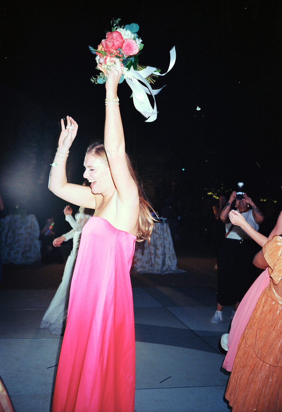 A woman in a pink strapless dress celebrating at night, holding a bouquet of flowers above her head with a big smile.