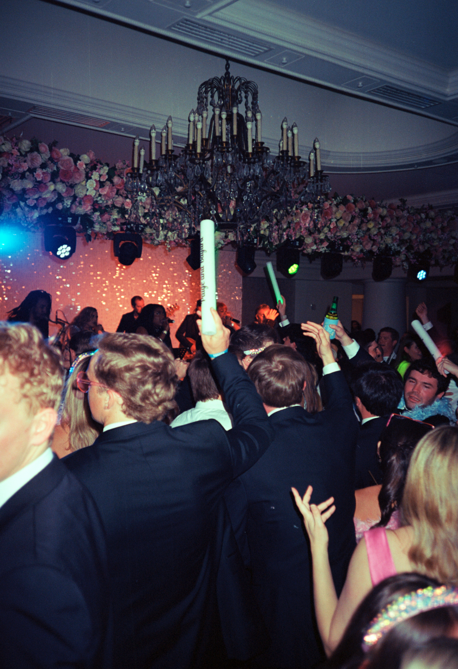Crowd of people at a lively party or celebration dance floor, many holding glow sticks or bottles, with a decorated ceiling and chandelier, and a band or musicians performing on stage in the background.