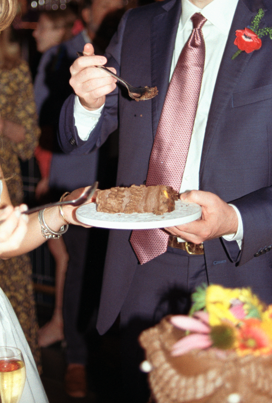 A man in a suit and tie holding a plate with a chocolate cake at a celebration or party, with people in the background.