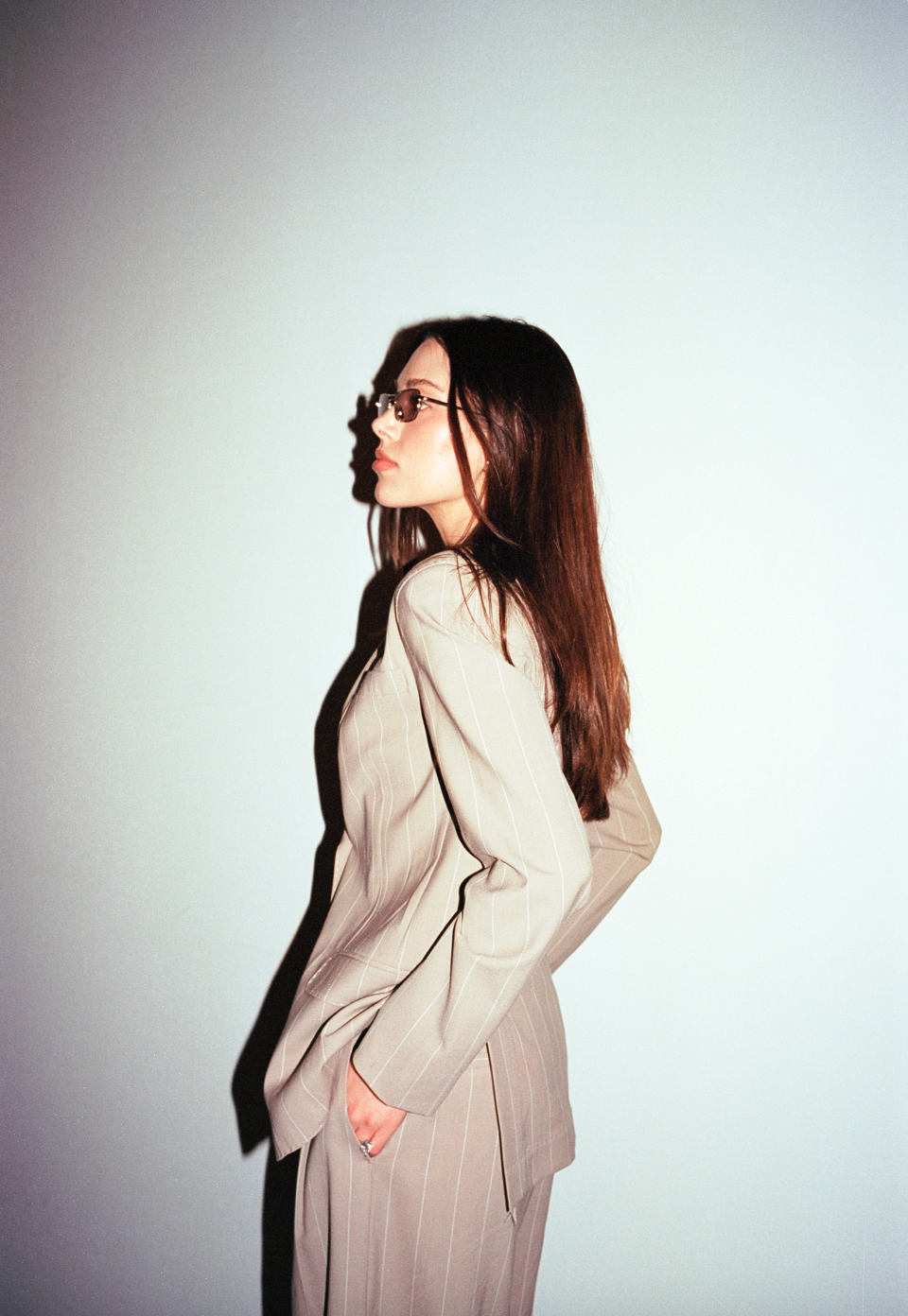 A woman in a beige pinstripe suit standing against a plain background, with her hands in her pockets, wearing glasses, and looking to the side.