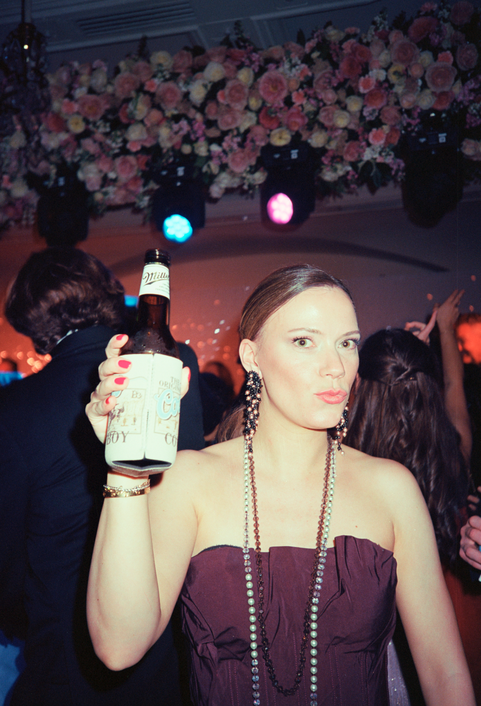A woman at a party holding a beer bottle, wearing a strapless purple dress and long beaded necklaces, with people dancing in the background and a floral decoration with colored lights overhead.