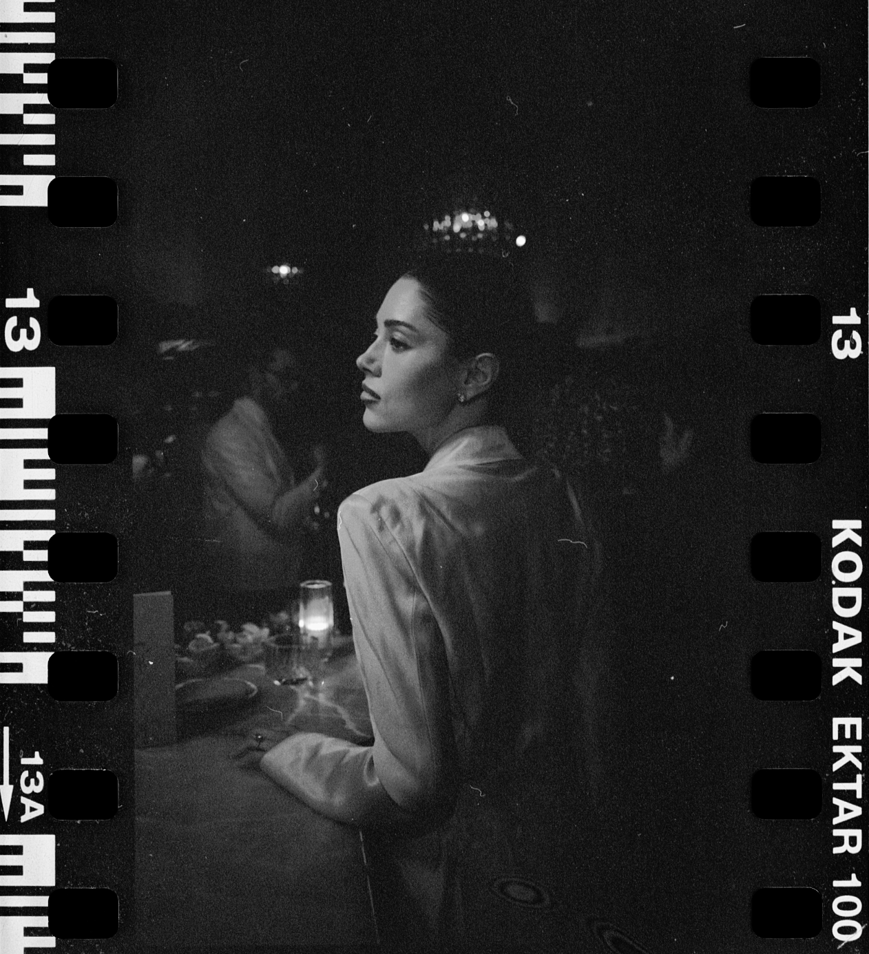 A black and white photo of a woman sitting at a table with a lit candle, dressed in a satin jacket, at a dimly lit event or restaurant, with other people and city lights in the background.