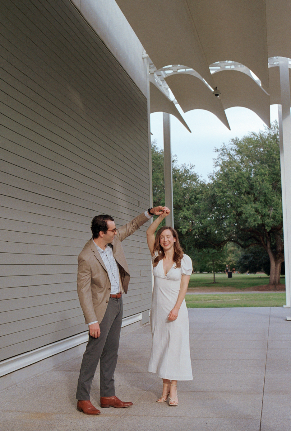 A man in a beige blazer and gray trousers dancing with a woman in a white dress outdoors, with trees and a modern building in the background.