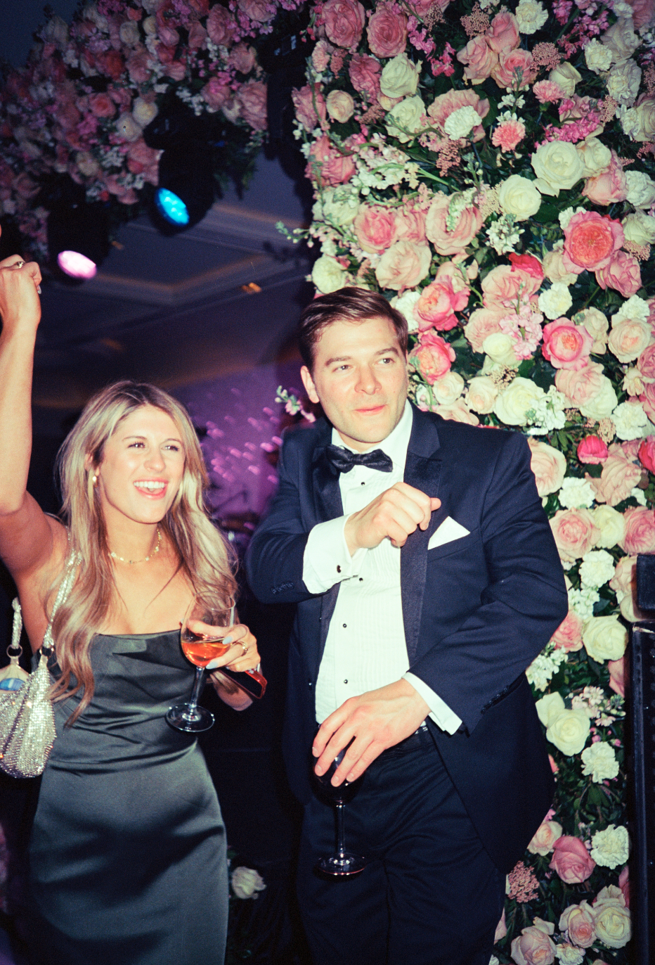 A man and woman dressed in formal attire, celebrating at an event with pink and white flower decorations in the background. The woman is holding a glass of wine, smiling, and raising her arm. The man is wearing a tuxedo with a bow tie, holding a wine