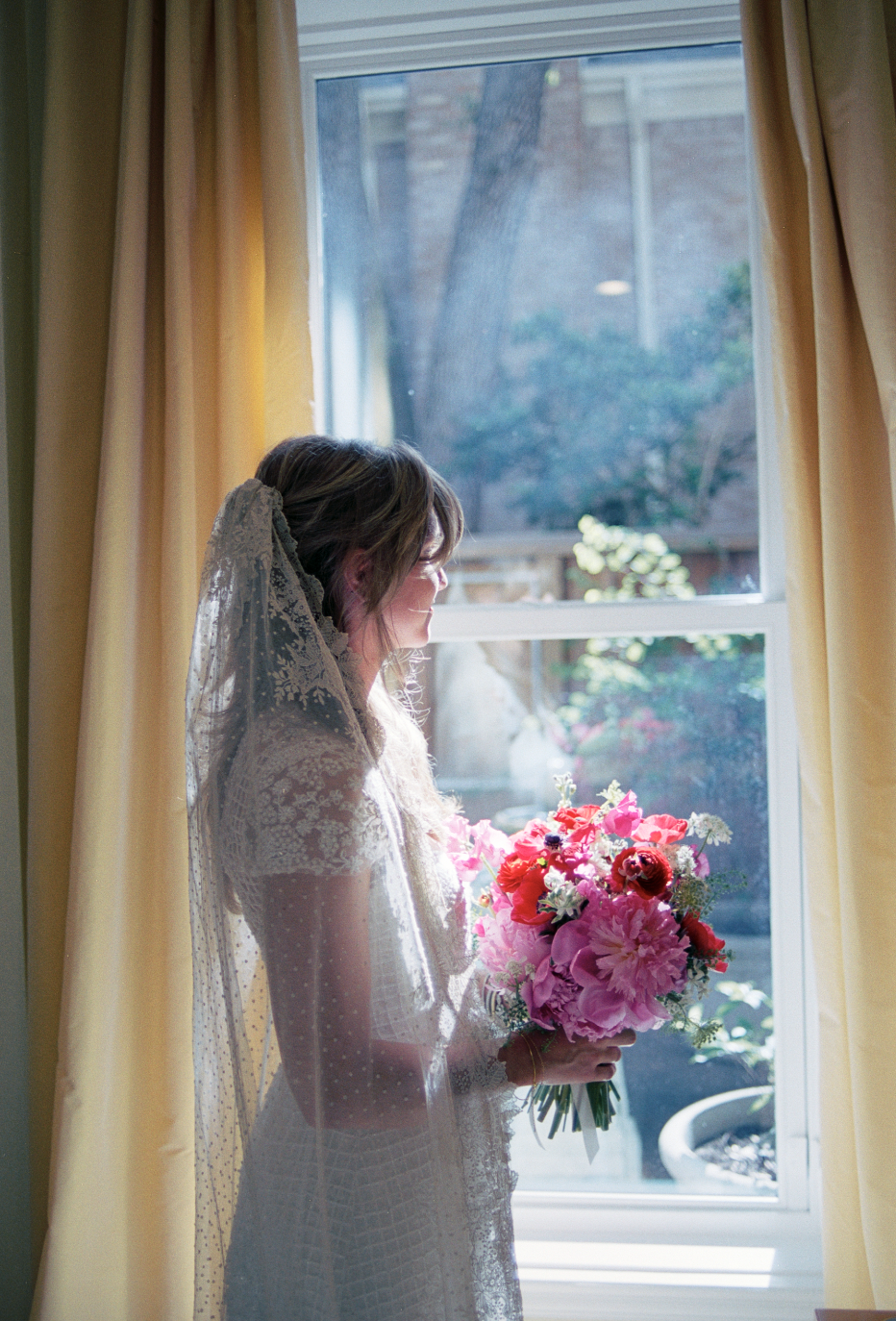A woman in a wedding dress, holding a bouquet of pink and red flowers, standing by a window with yellow curtains, with sunlight lighting her face and dress.