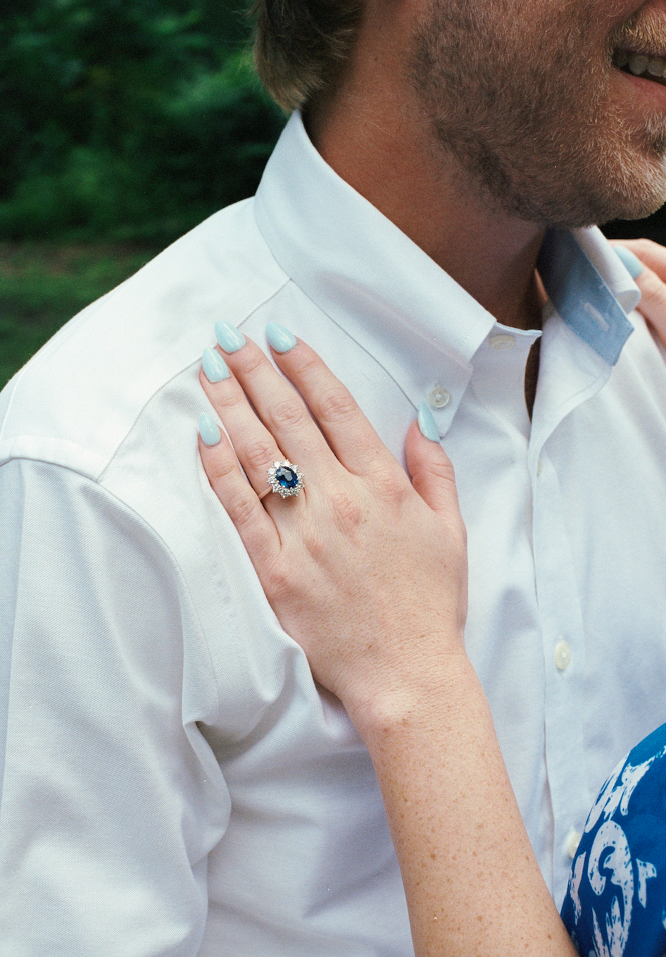 Close-up of a woman with light blue nails and a large blue gemstone ring resting on a man's shoulder, who is wearing a white dress shirt outdoors.