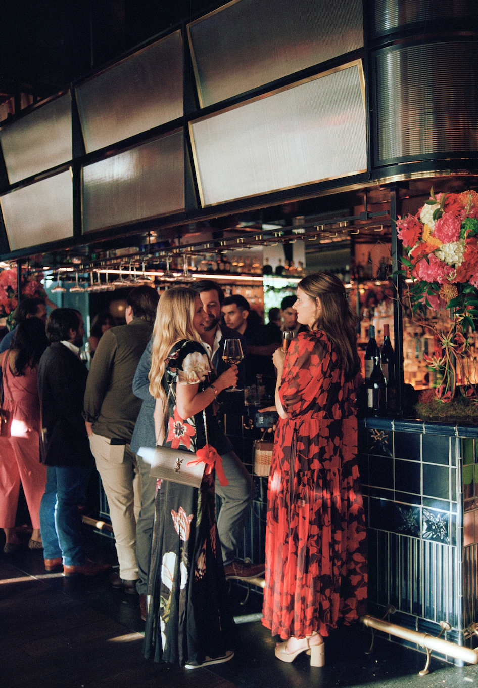 People socializing at a bar or lounge with floral arrangements and large display screens above.