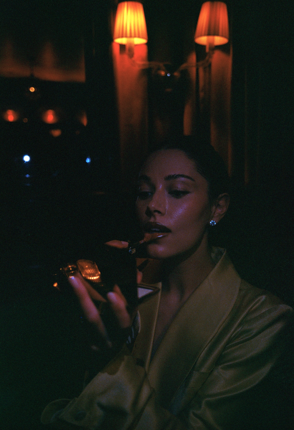 A woman in a satin jacket with makeup and earrings is in a dimly lit room, lighting a cigarette with a gold lighter, illuminated by warm orange lamps on the wall behind her.