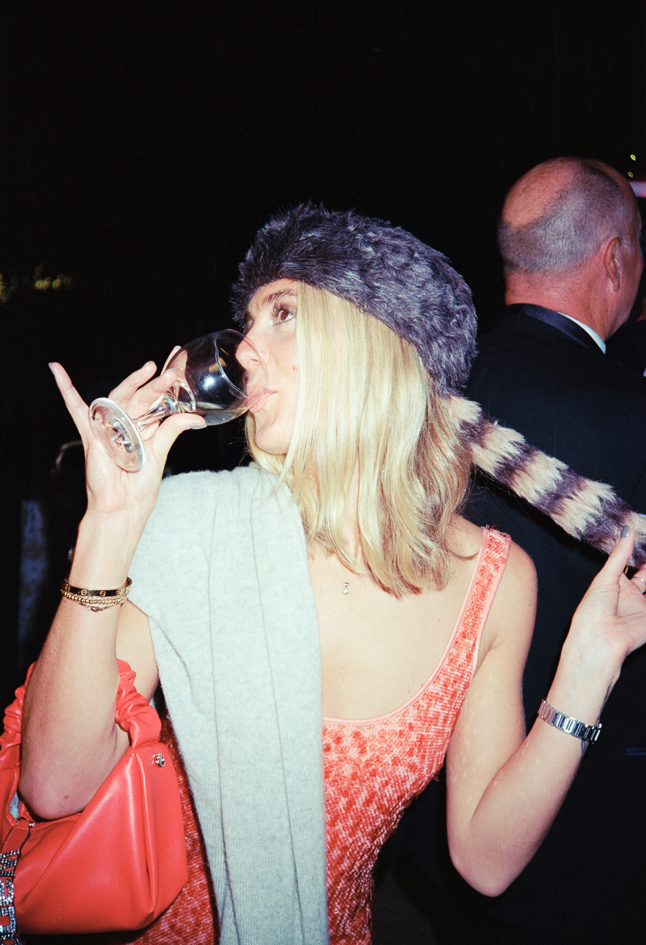 A woman drinking wine at a nighttime event, wearing a furry hat, a red dress, with a red handbag and a watch, holding a gray scarf, standing next to a man in a black suit.