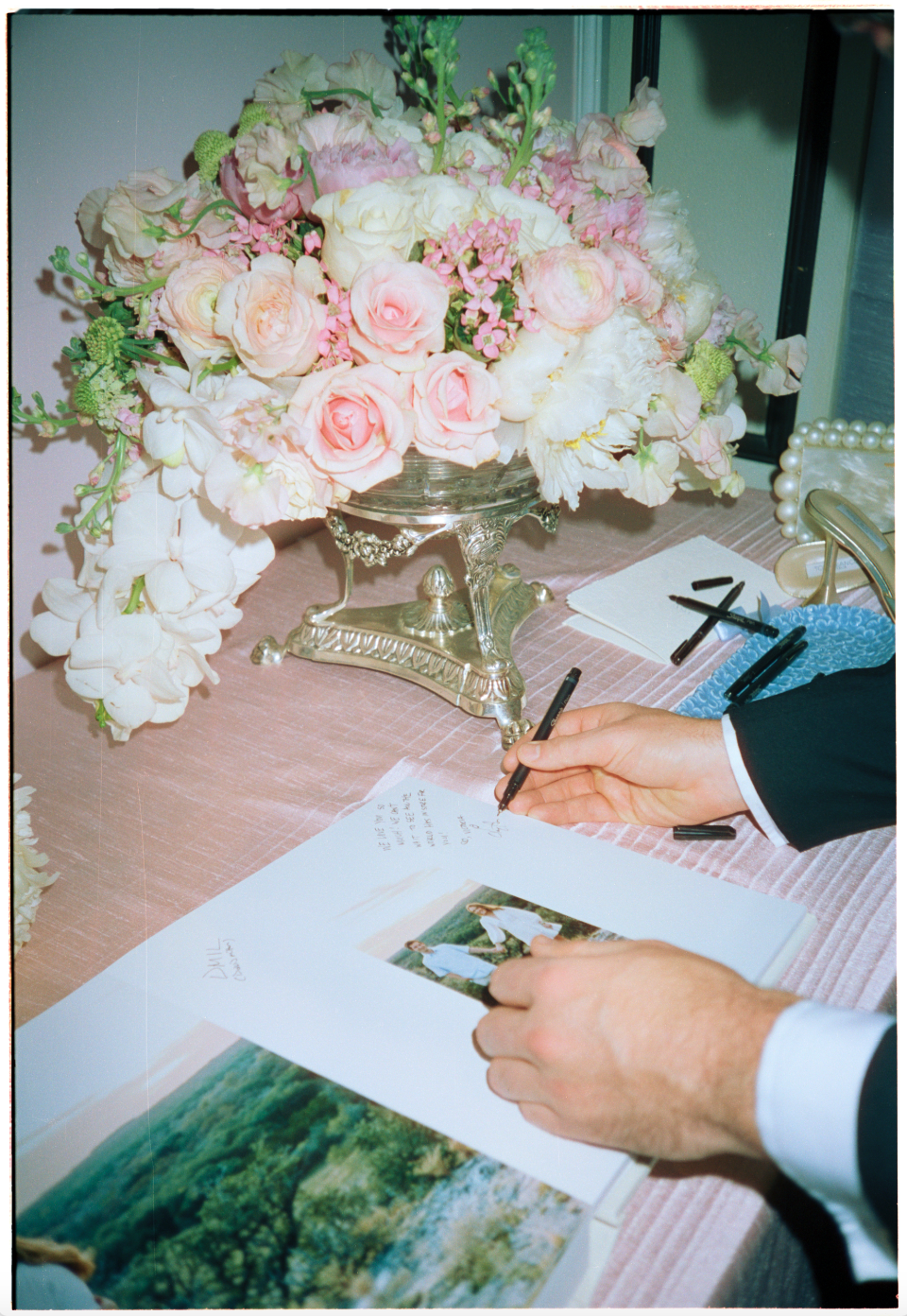A person signing a document or guestbook on a pink table with a large floral centerpiece and various writing supplies around.