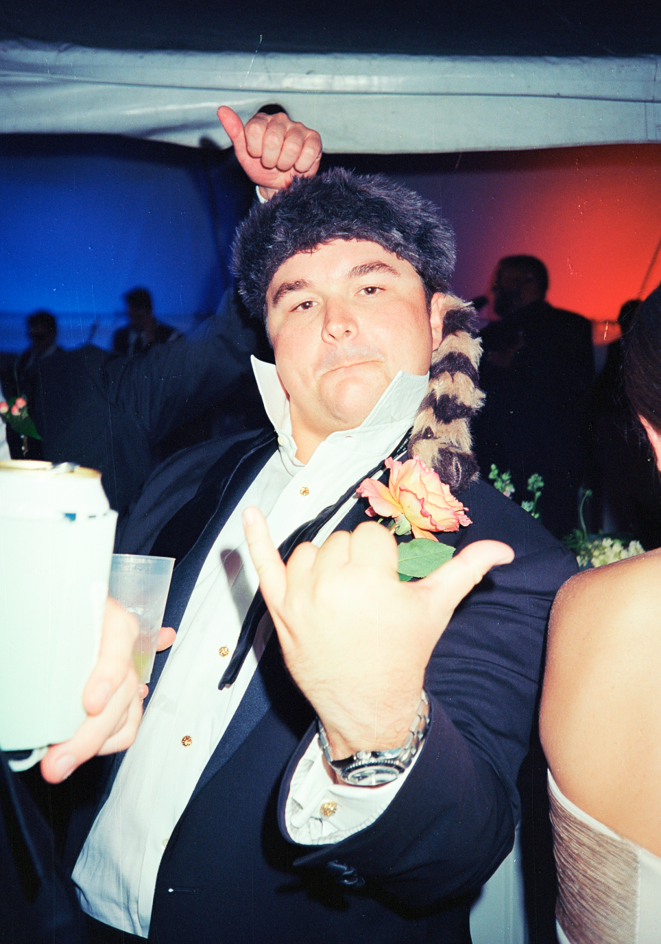 Man wearing a tuxedo and furry hat, making a shaka sign at a festive event, with a pink flower on his lapel and people in the background.