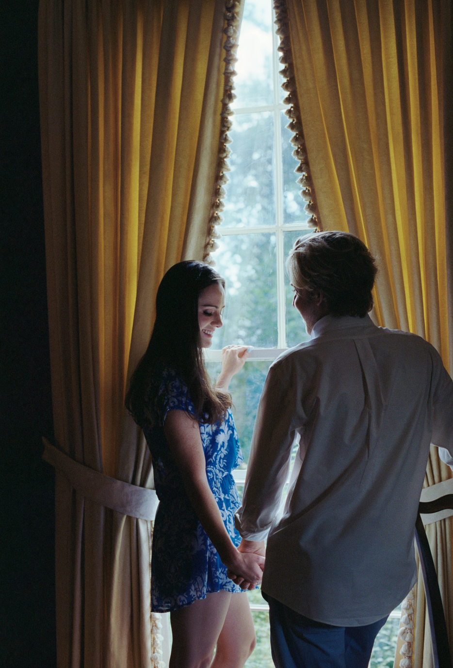 A young woman and a man holding hands in front of a window with yellow curtains, smiling at each other.