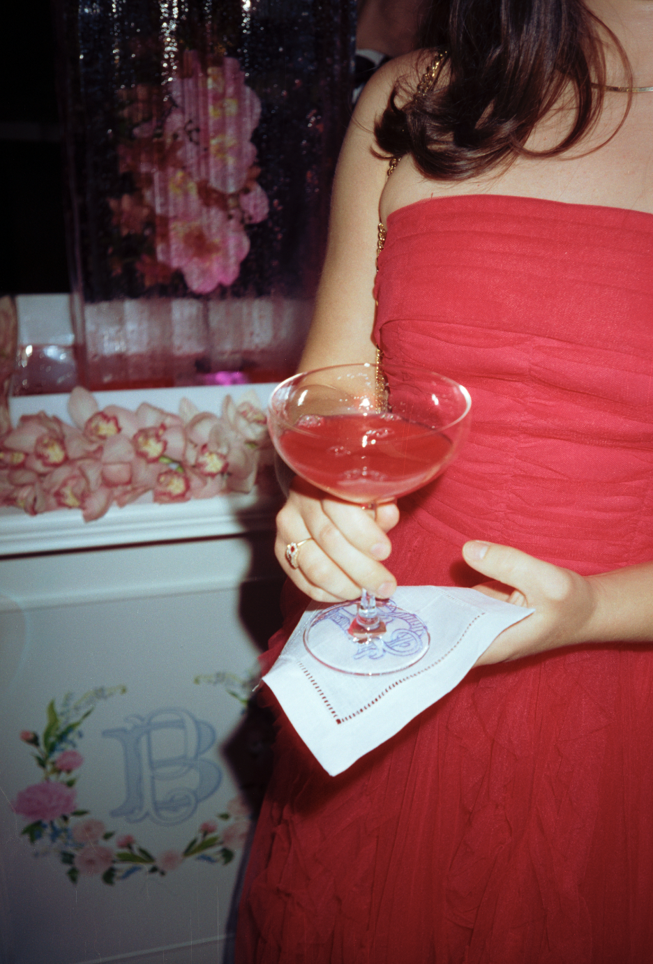 A woman in a red dress holding a cocktail glass with pink drink, resting her hand on a white napkin, at a celebration with pink flowers in the background.