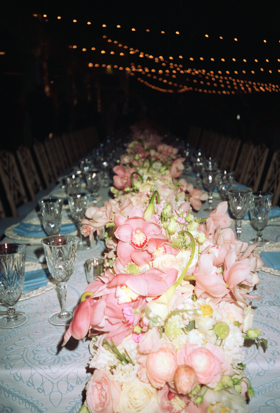 A long banquet table decorated with a pink floral centerpiece, set with wine glasses and blue napkins, under string lights at an evening event.