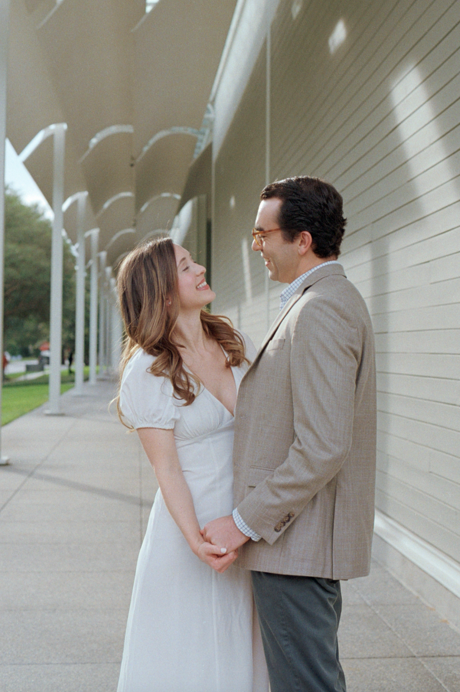 A couple standing close together, holding hands and smiling at each other outside, near a building with a striped wall and a row of white arches.